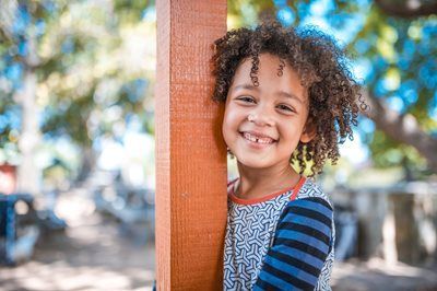 Smiling child peeks from behind a wooden post in a sunlit park, with blurred trees in the background