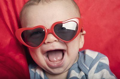 A baby wearing large, red heart-shaped sunglasses, laughing against a solid red background.