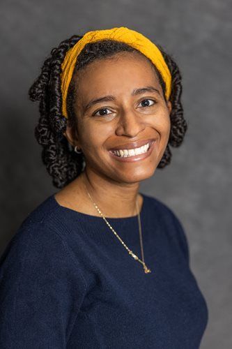 Smiling person with curly hair and yellow headband, wearing a navy top against a gray background