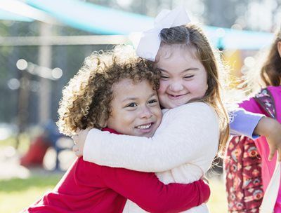 Two children hugging outdoors, one in a red sweater and one in a white sweater, both smiling