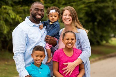 Family of five smiling outdoors, with parents holding three children on a path.