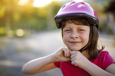 Smiling child wearing a purple helmet, adjusting chin strap outdoors on a sunny day