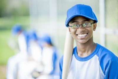 Smiling boy in blue cap and glasses holding a baseball bat on a field