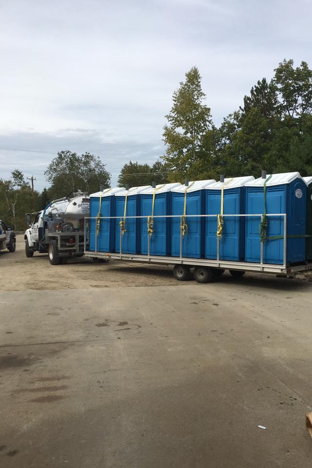 A truck towing a trailer loaded with multiple blue portable toilets. Outdoor setting, cloudy sky.