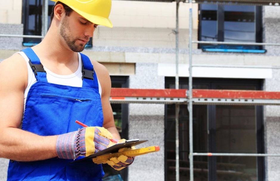 A man wearing a hard hat and overalls is writing on a clipboard.