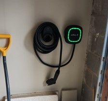 Wall-mounted EV charger with coiled black cable and green-lit display on a beige wall