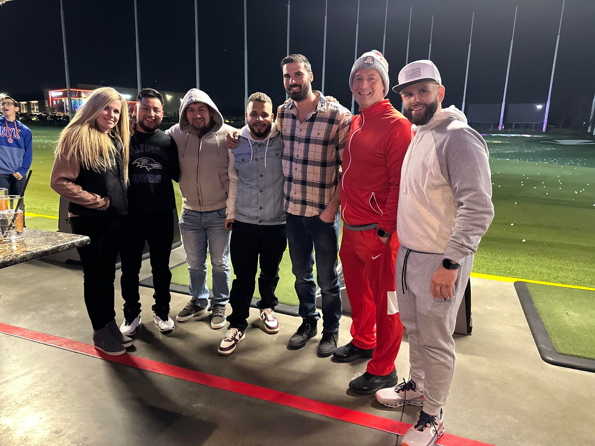 Six people posing together at a nighttime golf driving range, wearing casual and athletic clothes.