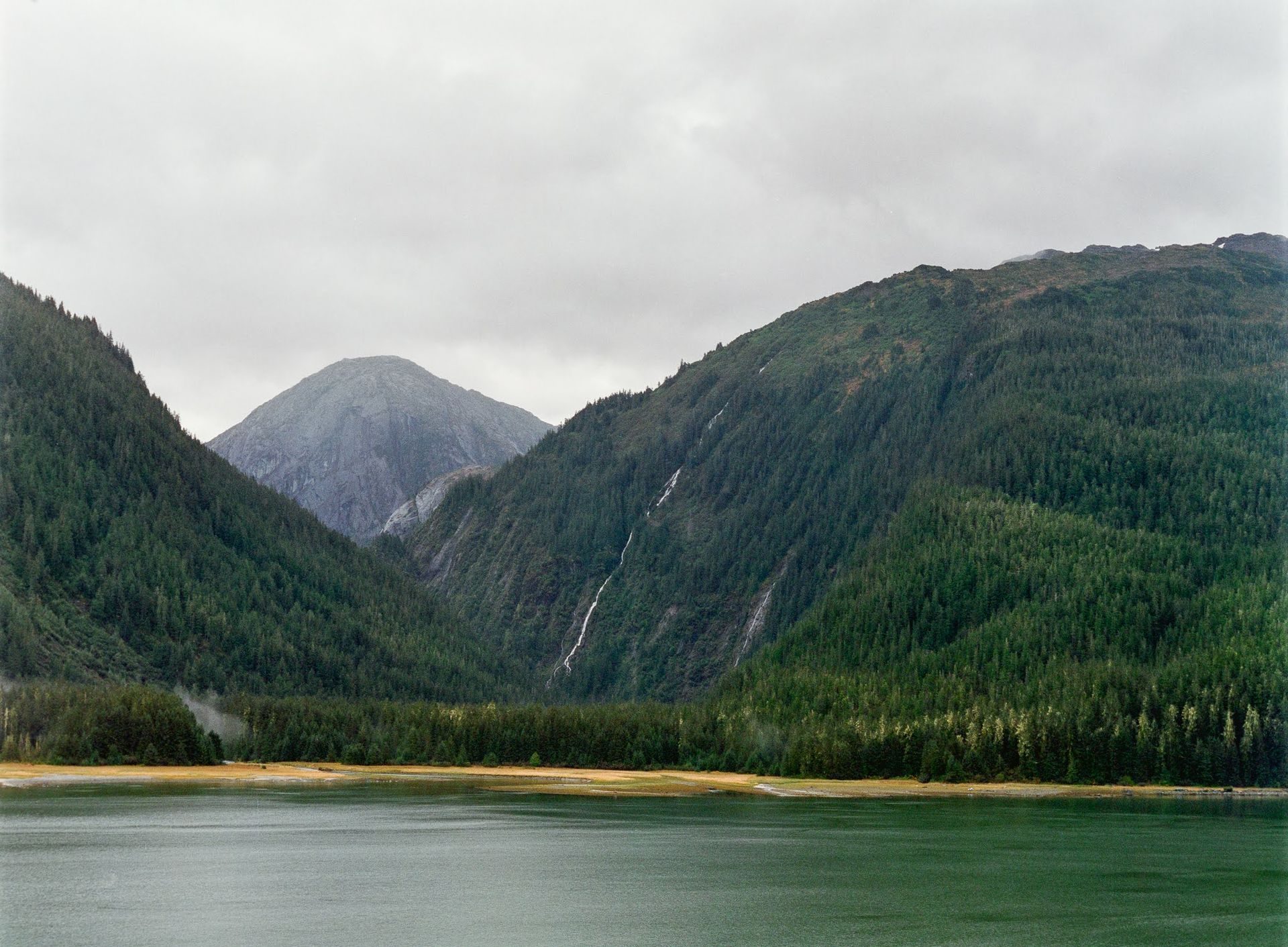 Cloudy mountain lake with forested hills and a distant peak reflected in calm water Cloudy mountain lake with forested hills and a distant peak reflected in calm water