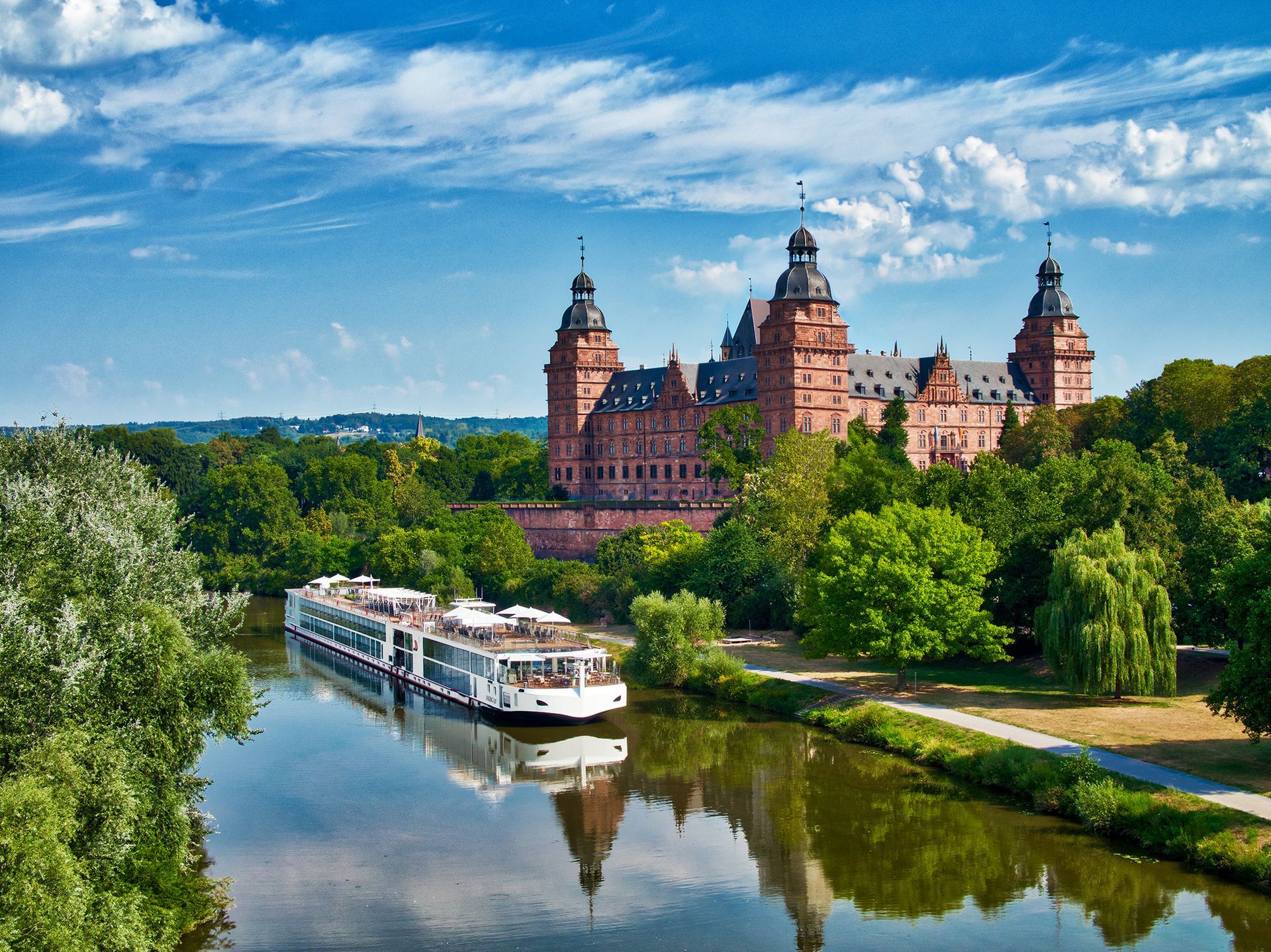 A large castle with multiple towers sits by a river with a cruise ship, under a blue sky.