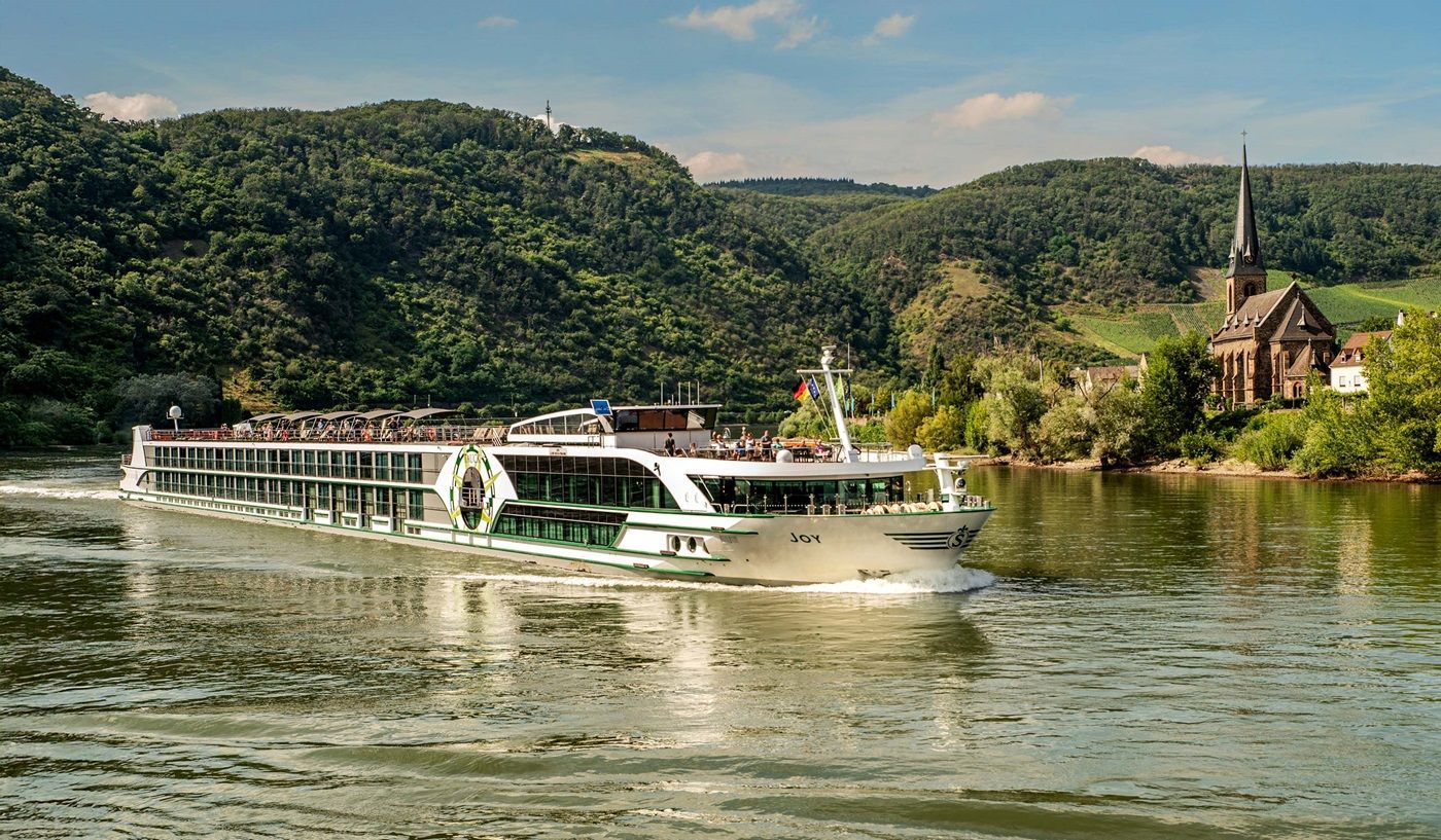 River cruise ship sailing on water, lush green hills in the background with a church.