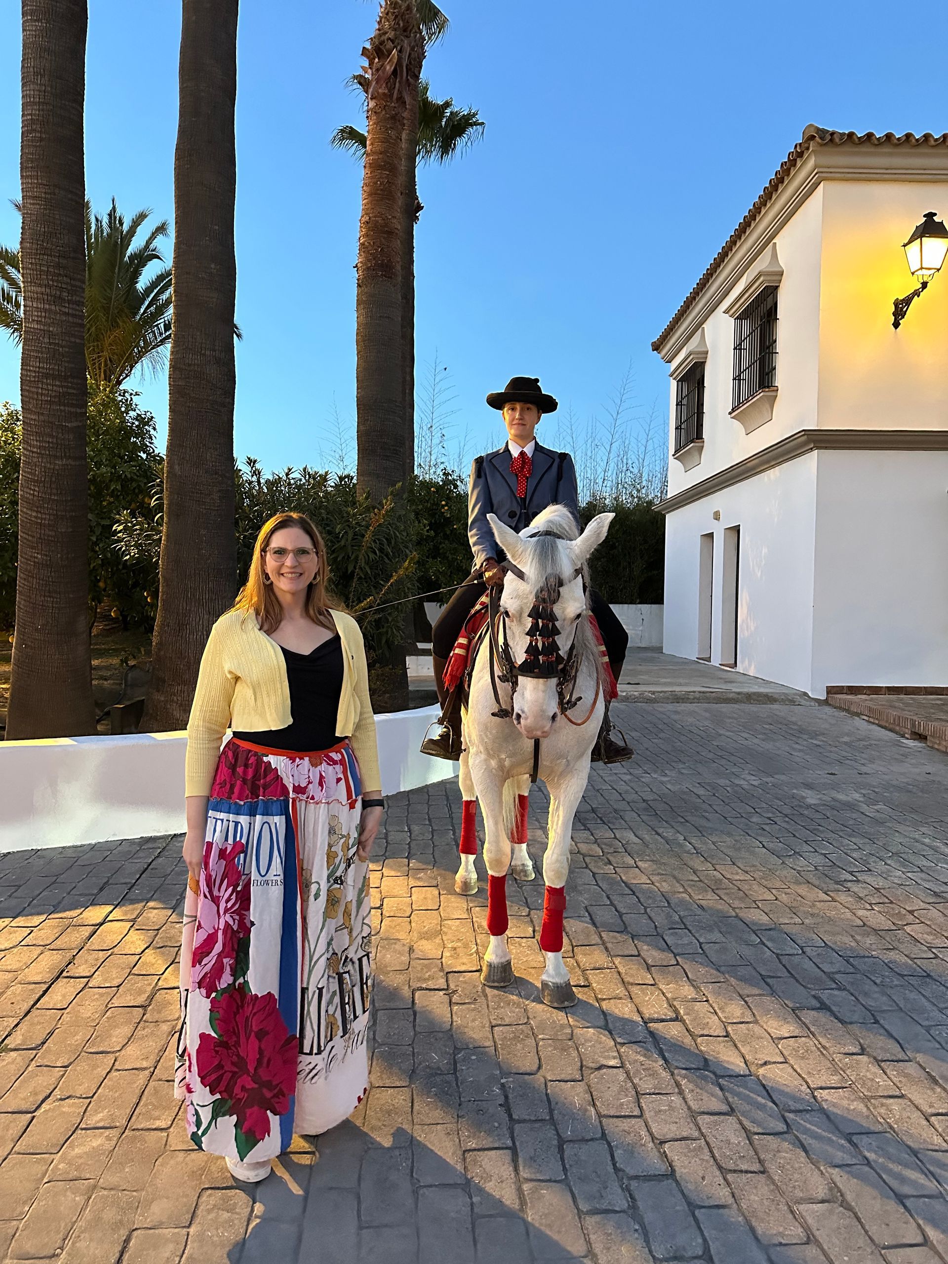 Woman beside a rider on a white horse, both in formal wear, in front of a white building with palm trees. Woman beside a rider on a white horse, both in formal wear, in front of a white building with palm trees.