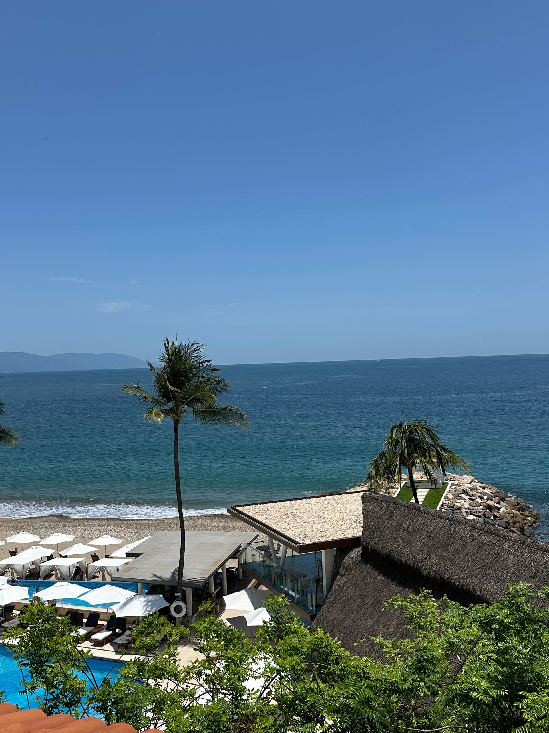 Beachfront resort with white umbrellas, blue pool, thatched roofs, and a calm blue ocean under a clear sky