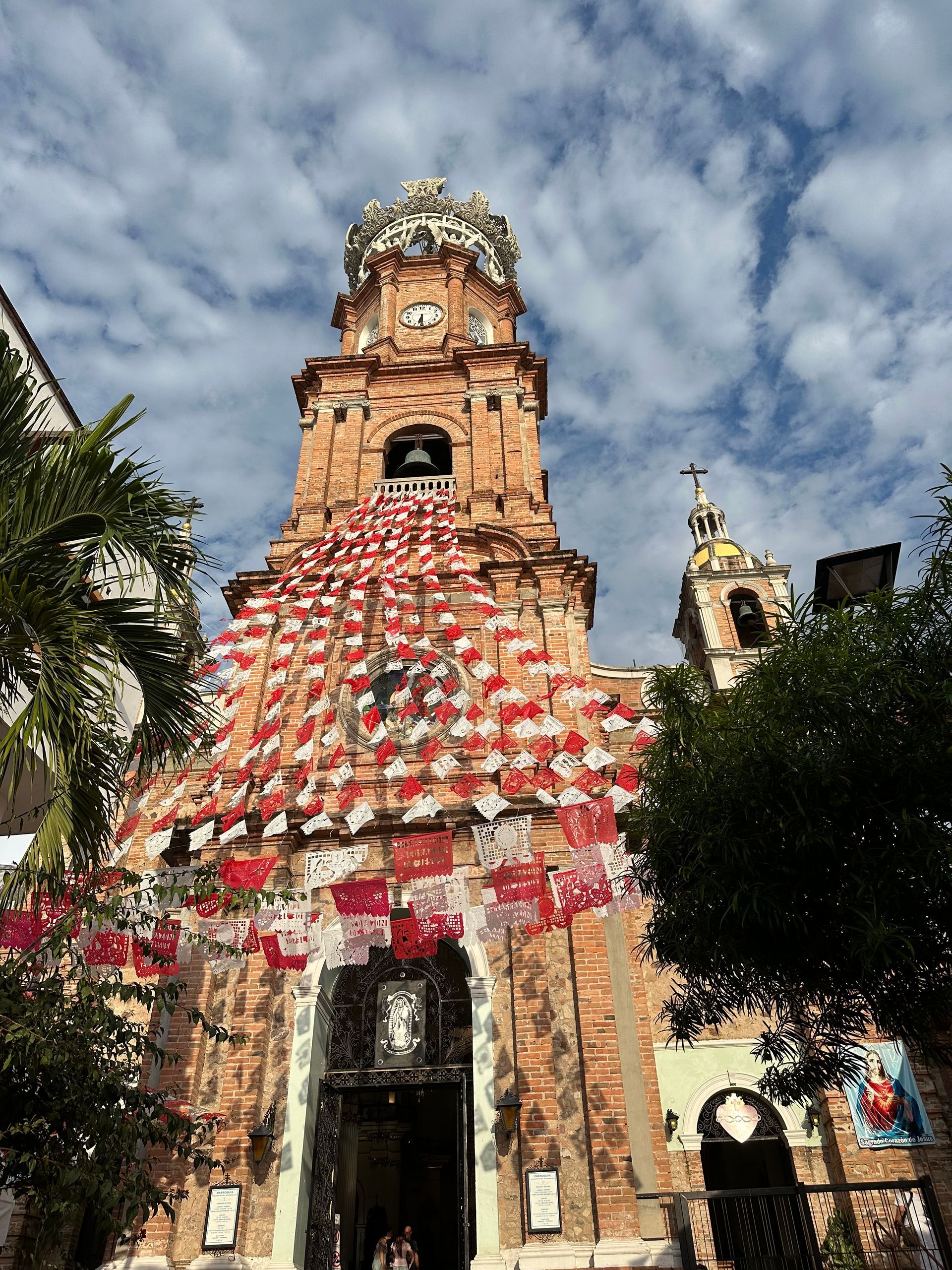 Historic church tower draped in red-and-white flags, viewed from below against a cloudy sky