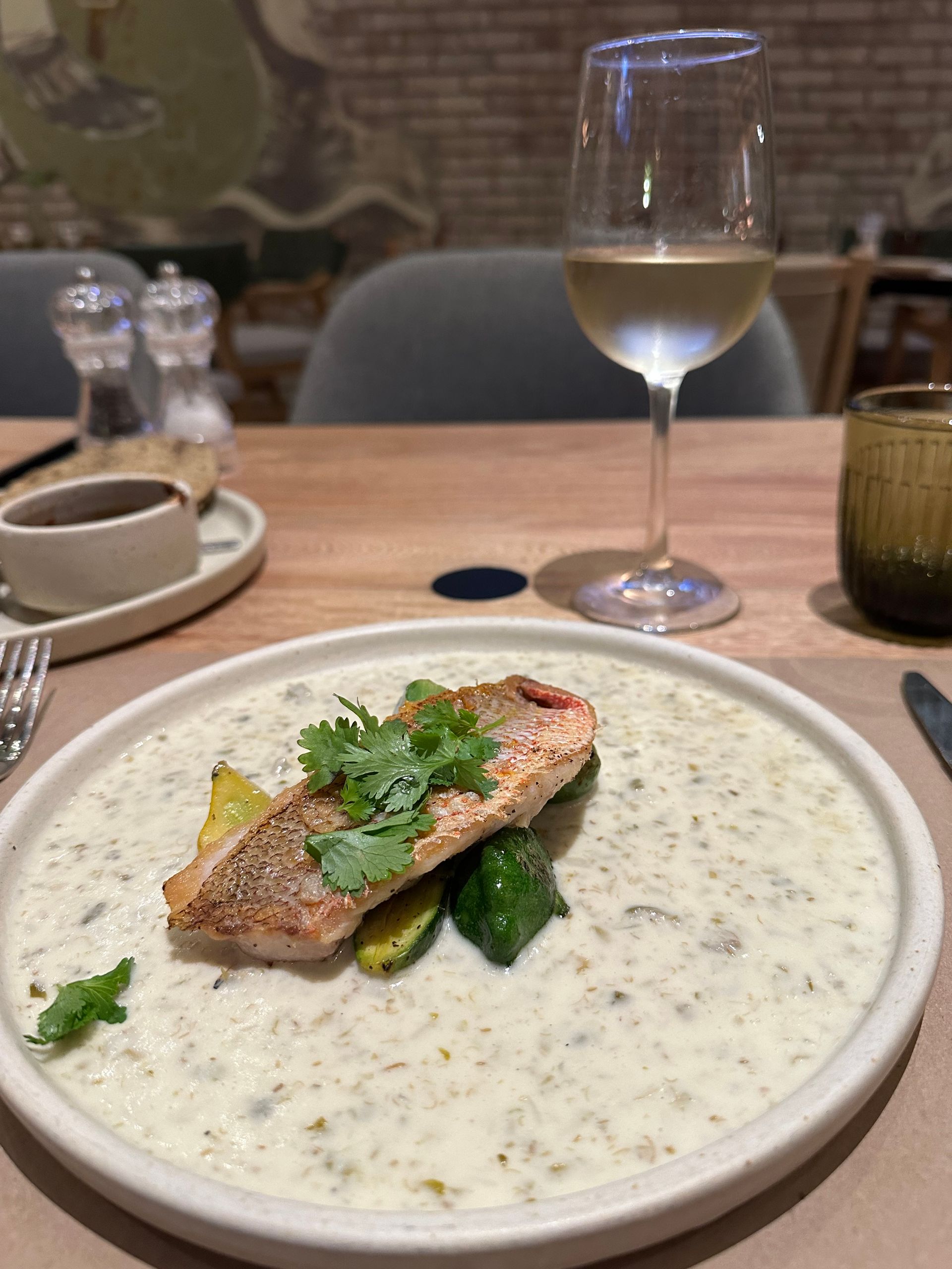 Restaurant plate of creamy soup with seared fish, on a table beside a glass of white wine.