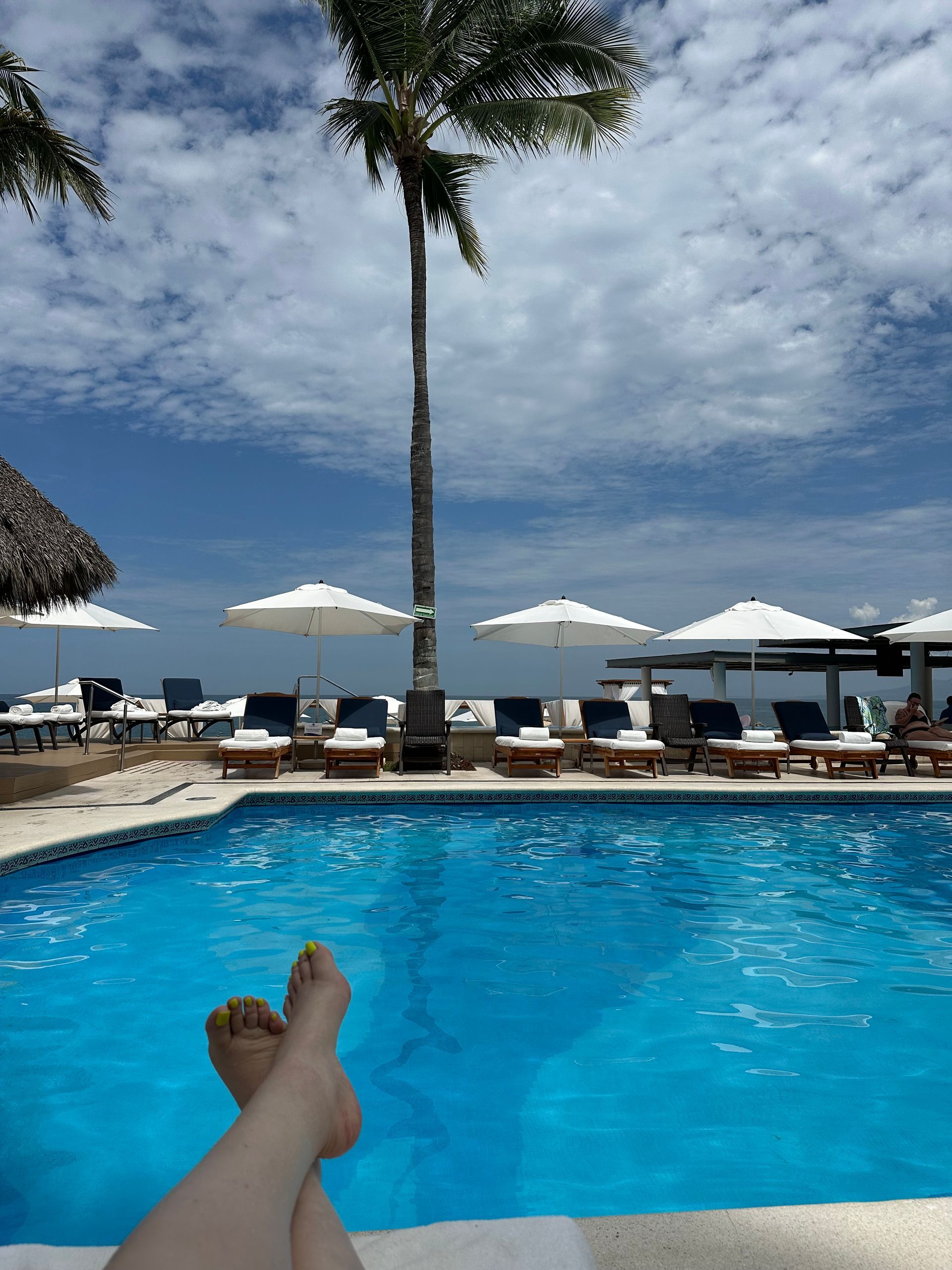 Poolside beach view with a person’s legs in the foreground, lounge chairs, umbrellas, and ocean beyond.