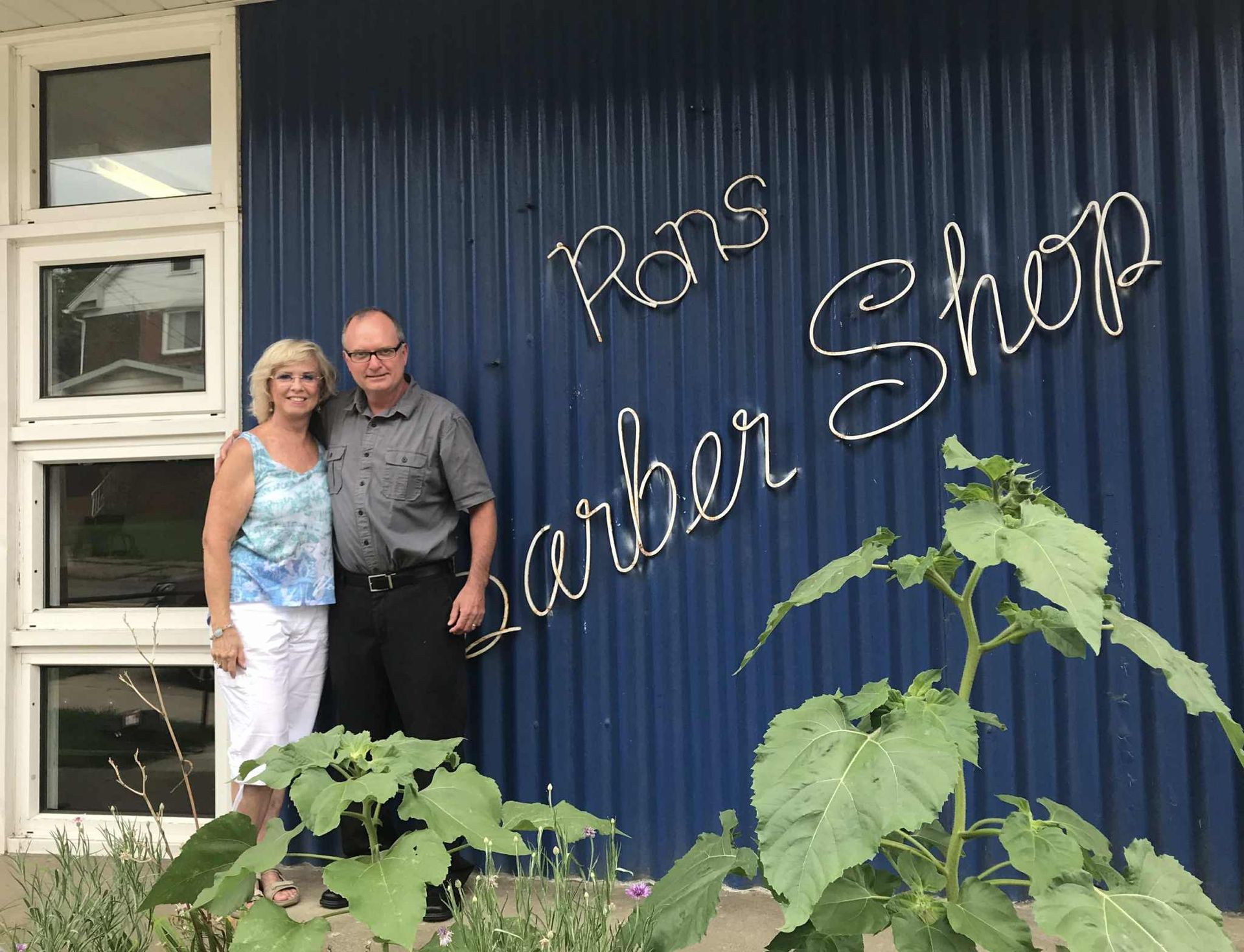 Couple standing in front of Ron's Barber Shop with blue corrugated metal wall and neon sign.