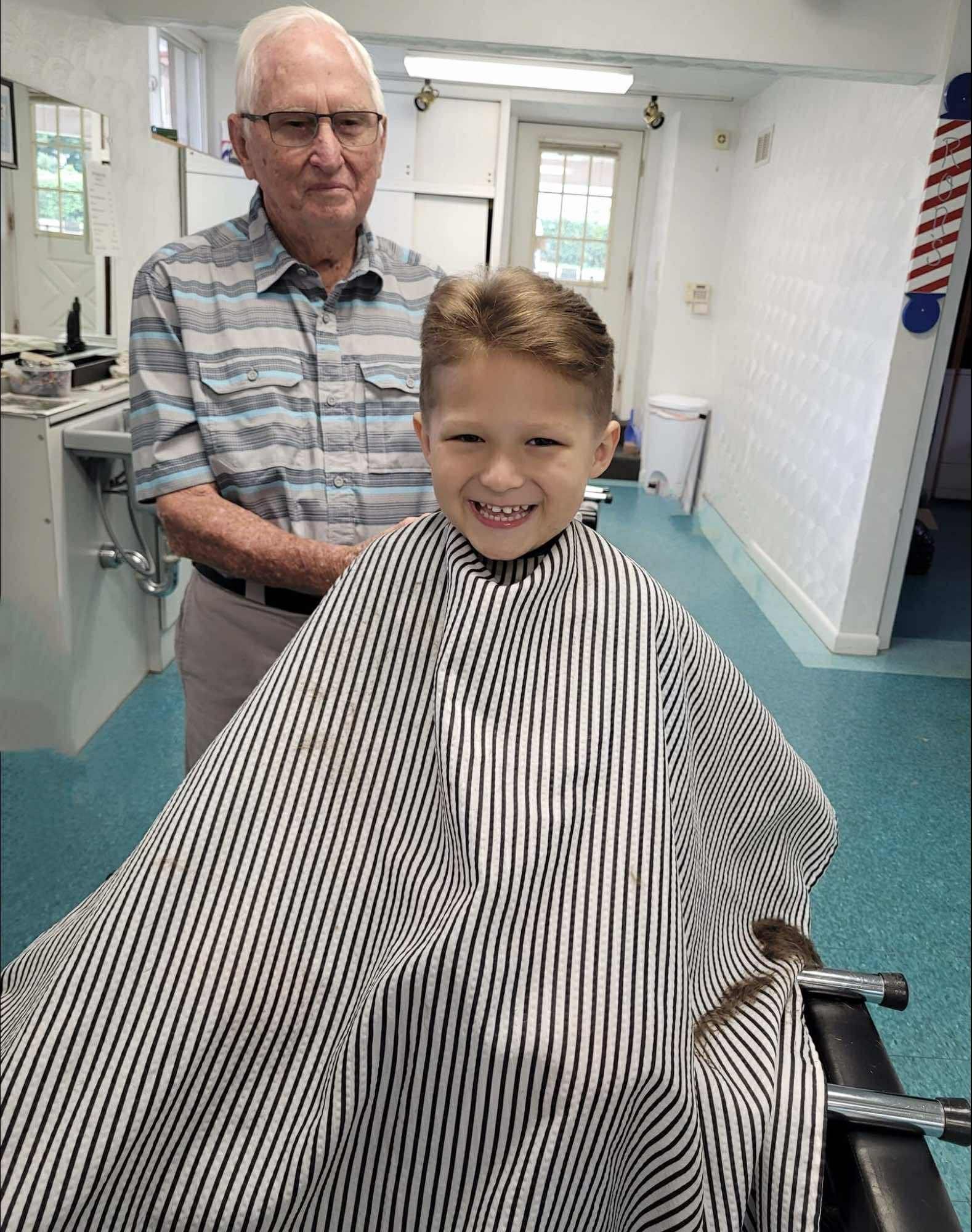 A smiling young boy in a barber cape getting a haircut from an older barber in a shop.