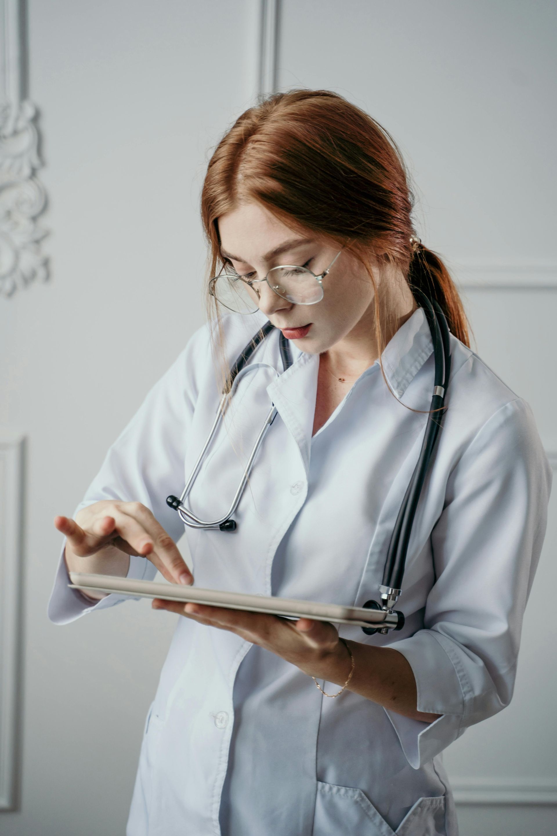 Woman doctor with red hair, wearing glasses, a white coat, and a stethoscope, using a tablet.