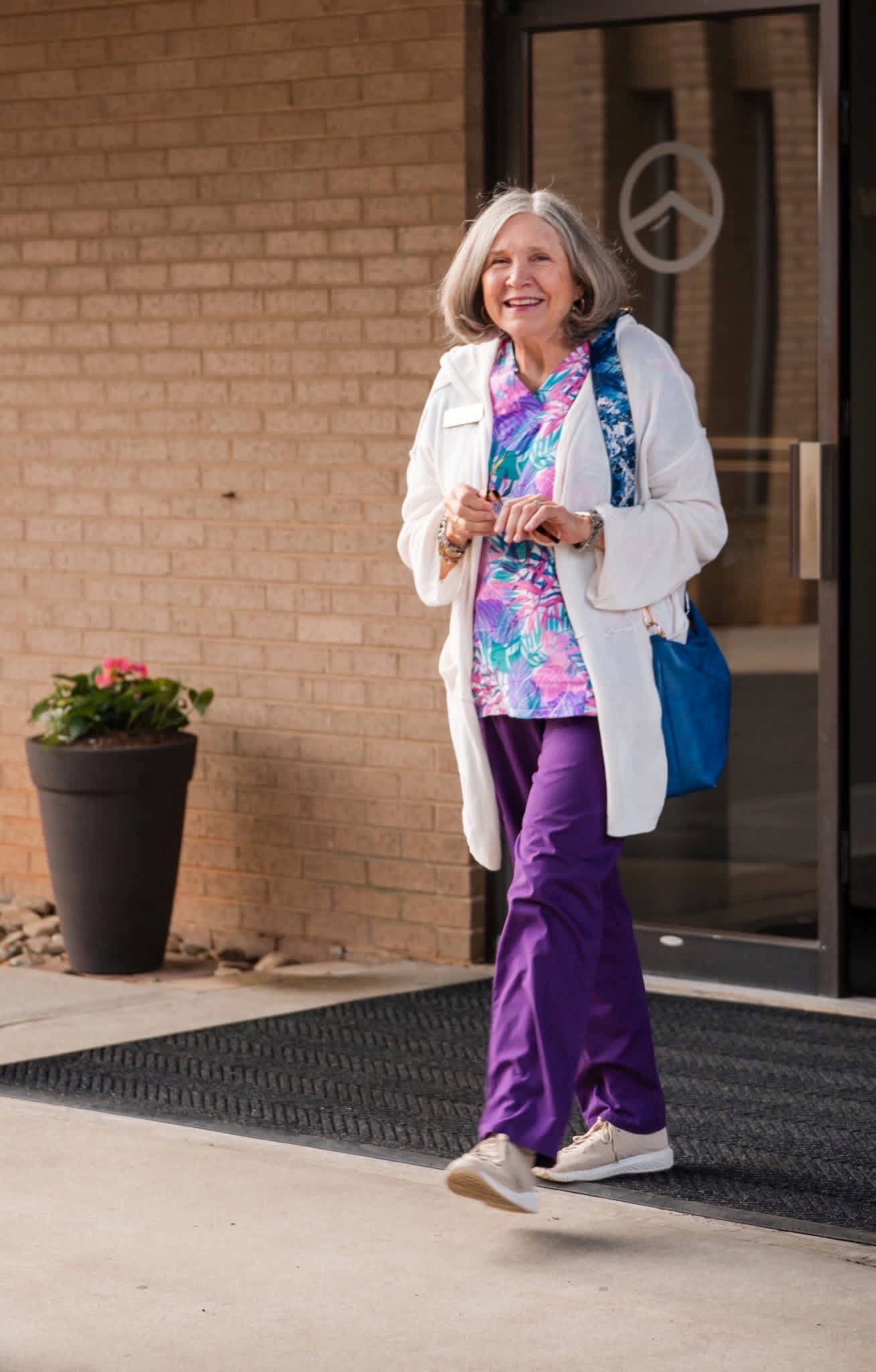 Smiling woman in white jacket and purple pants walking out of a building, holding a phone