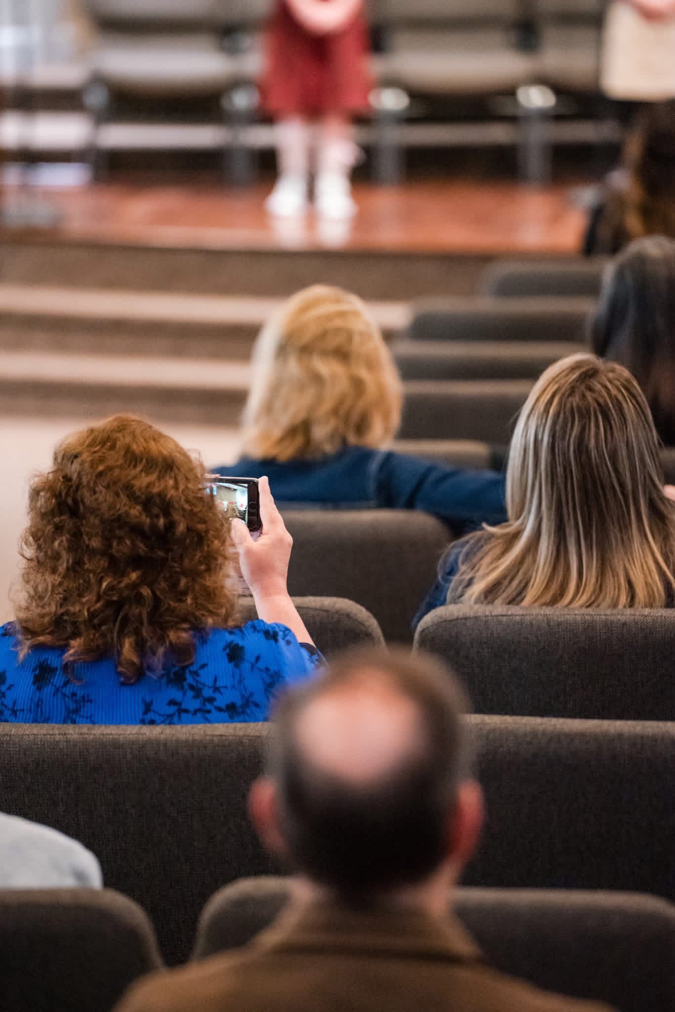 Audience seated in a theater, one person holding up a phone toward the stage