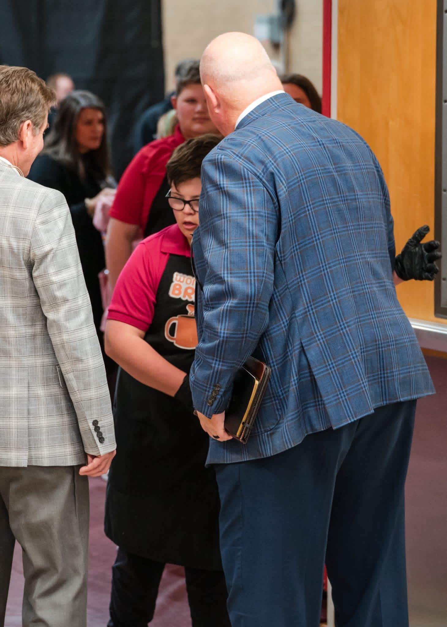 People chatting in a hallway; a man in a blue jacket stands beside a woman in a pink top holding a folder.