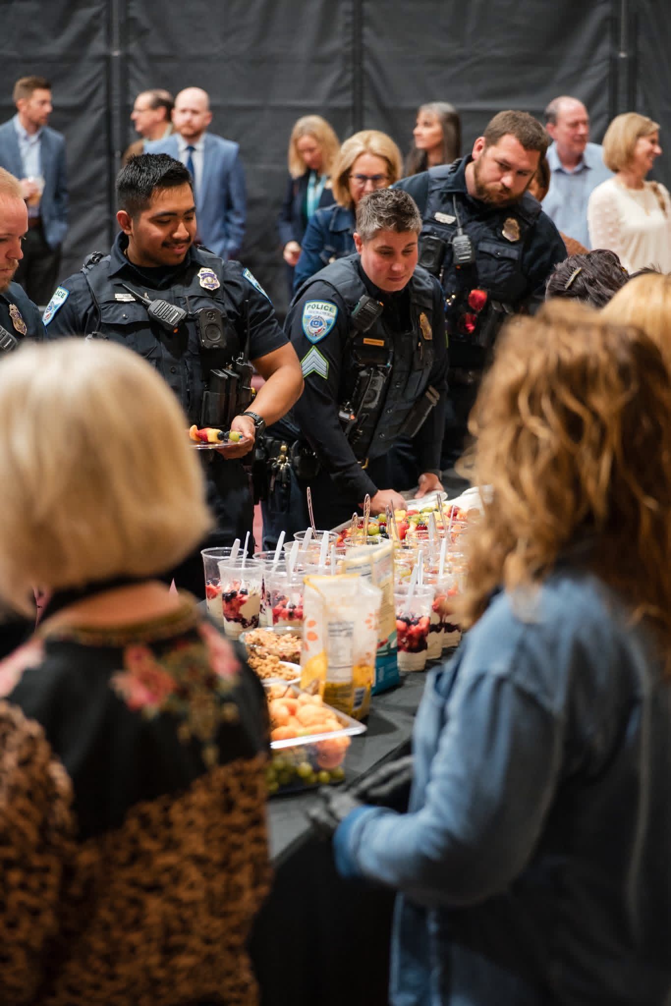 People in dark uniforms serving drinks at a crowded indoor event with a long table of colorful snacks