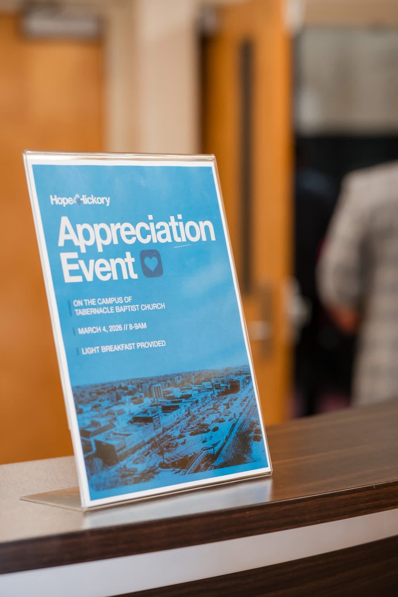 Blue “Appreciation Event” sign on a table in a lobby, with blurred people in the background