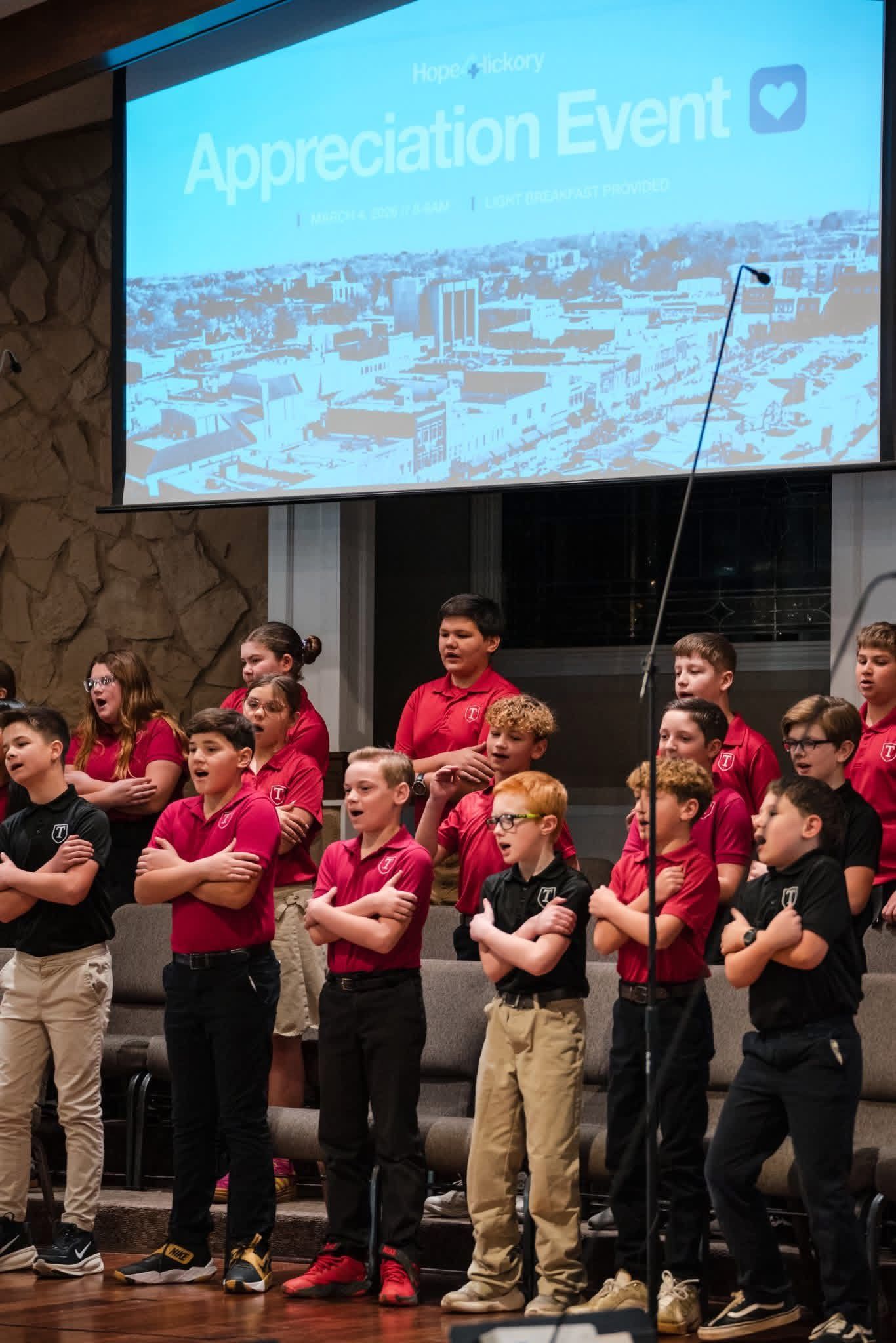 Choir in red and black shirts stands on stage beneath an “Appreciation Event” screen