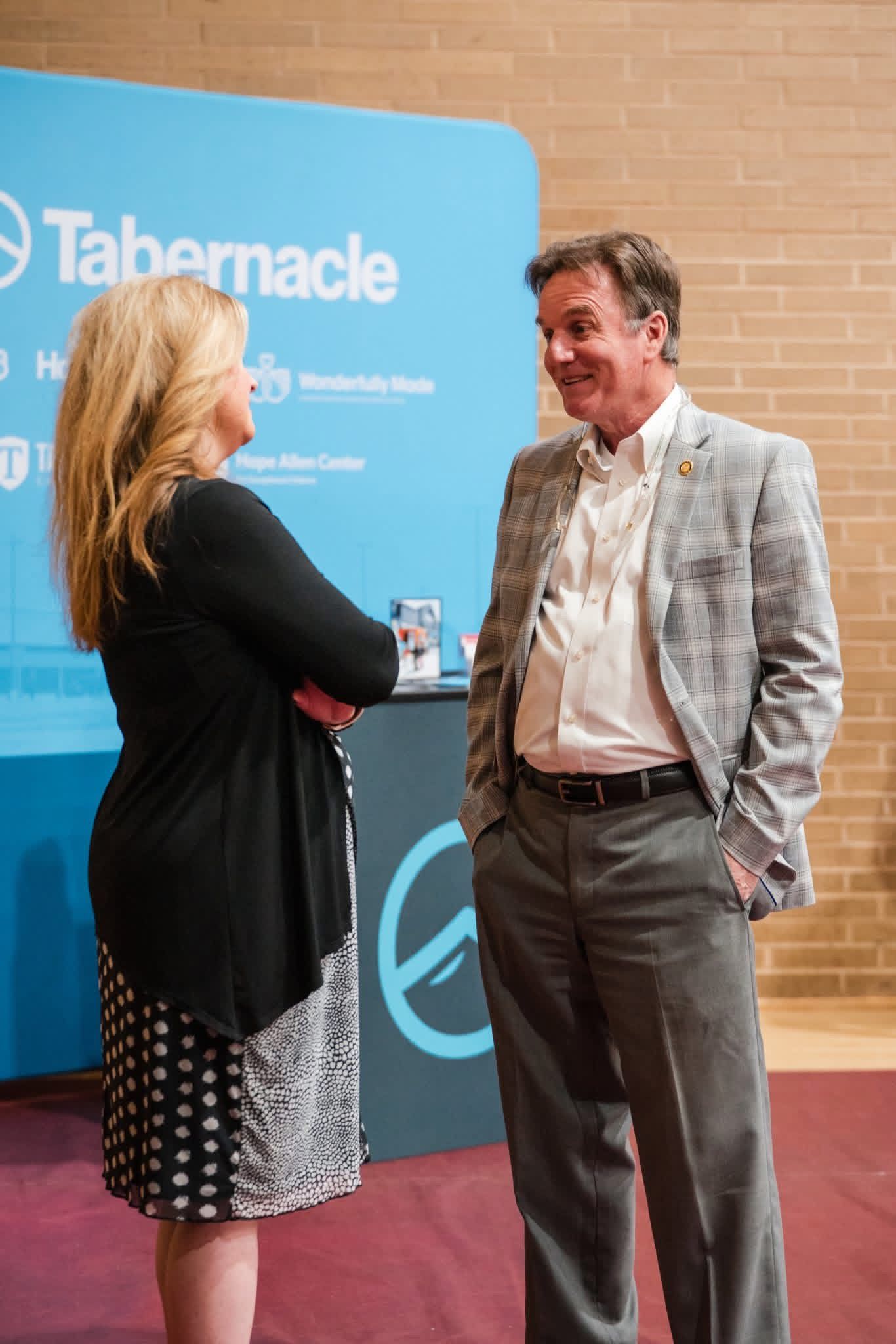 Two people talking at a Tabernacle event, with a blue backdrop and brick wall behind them.