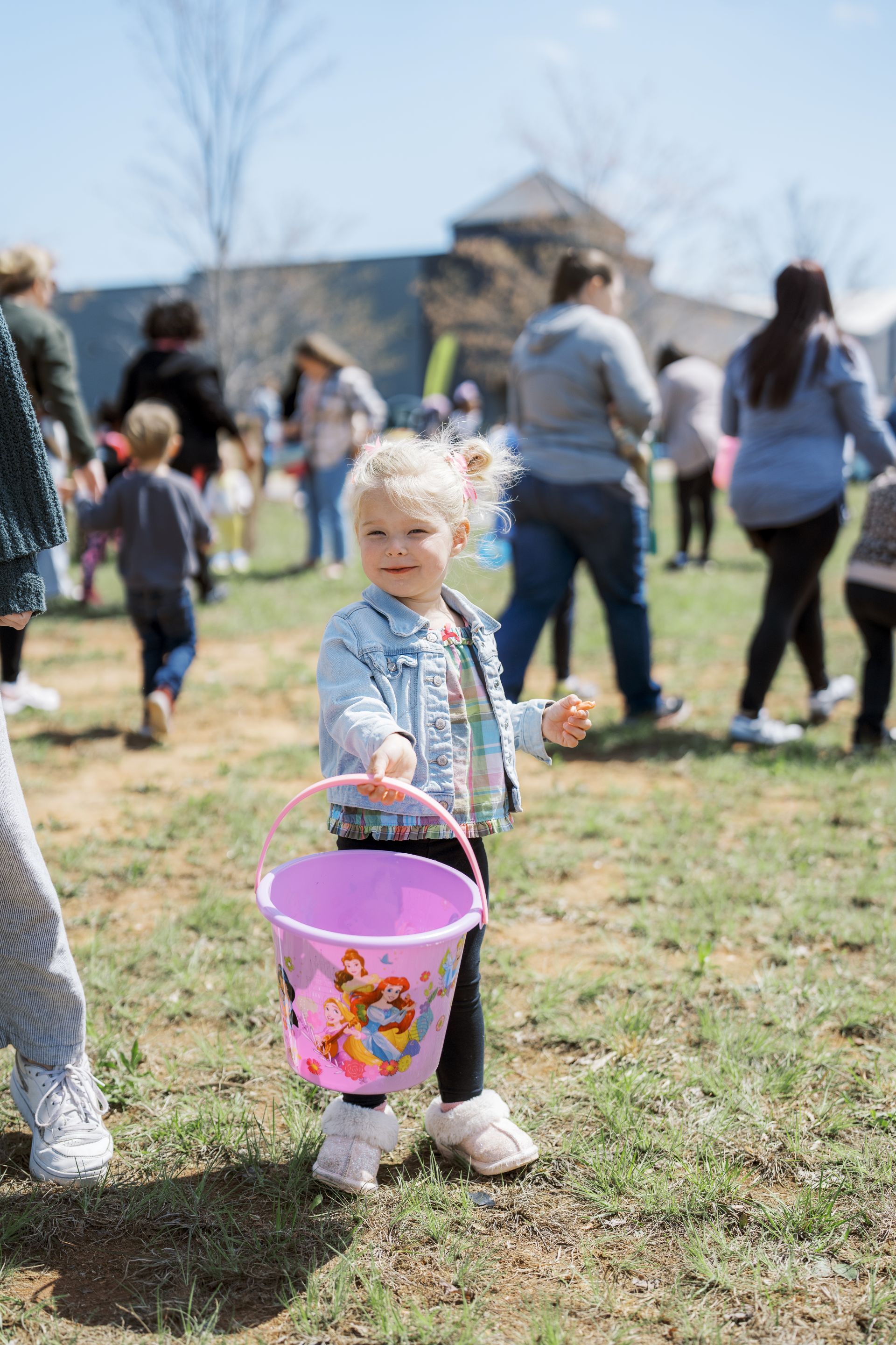 Child holding a pink Easter bucket in a grassy field during an outdoor egg hunt