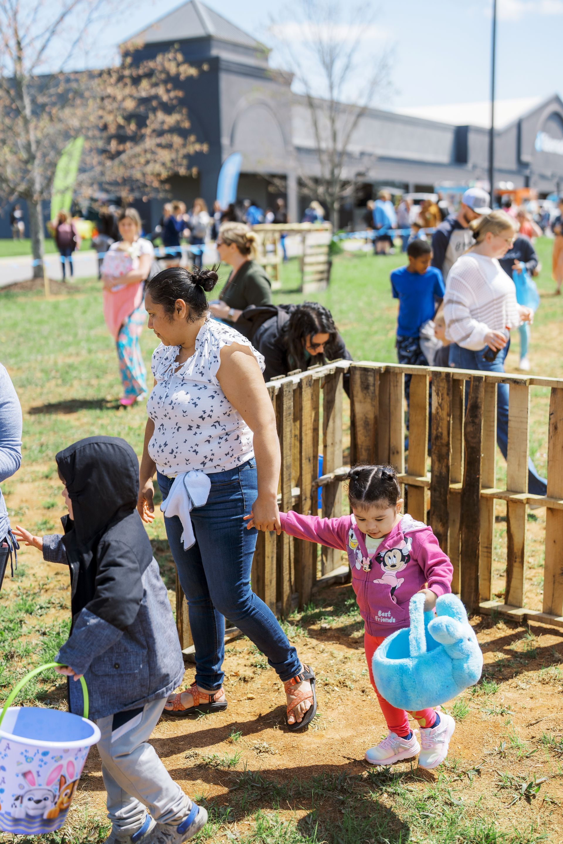 Children carrying Easter baskets walk through a sunny outdoor event near a wooden fence.