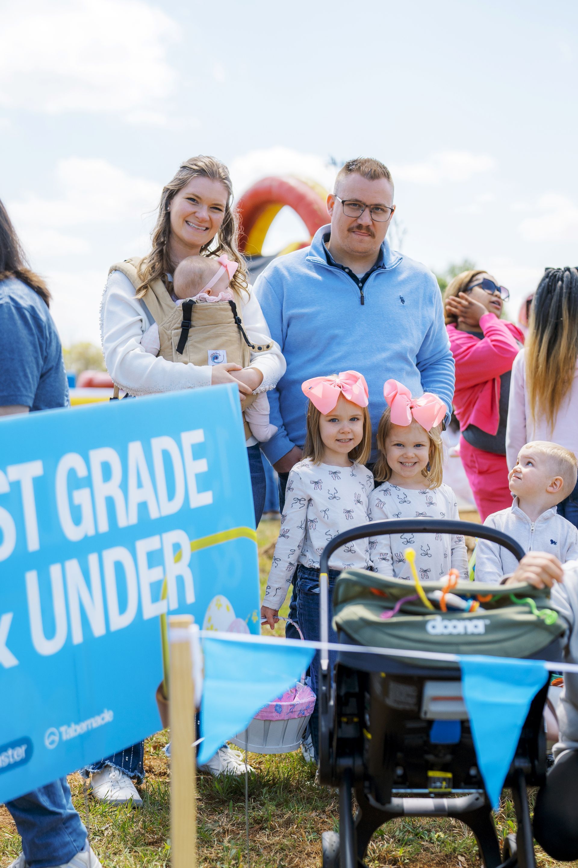Family standing outdoors at a community event beside a blue sign and a stroller