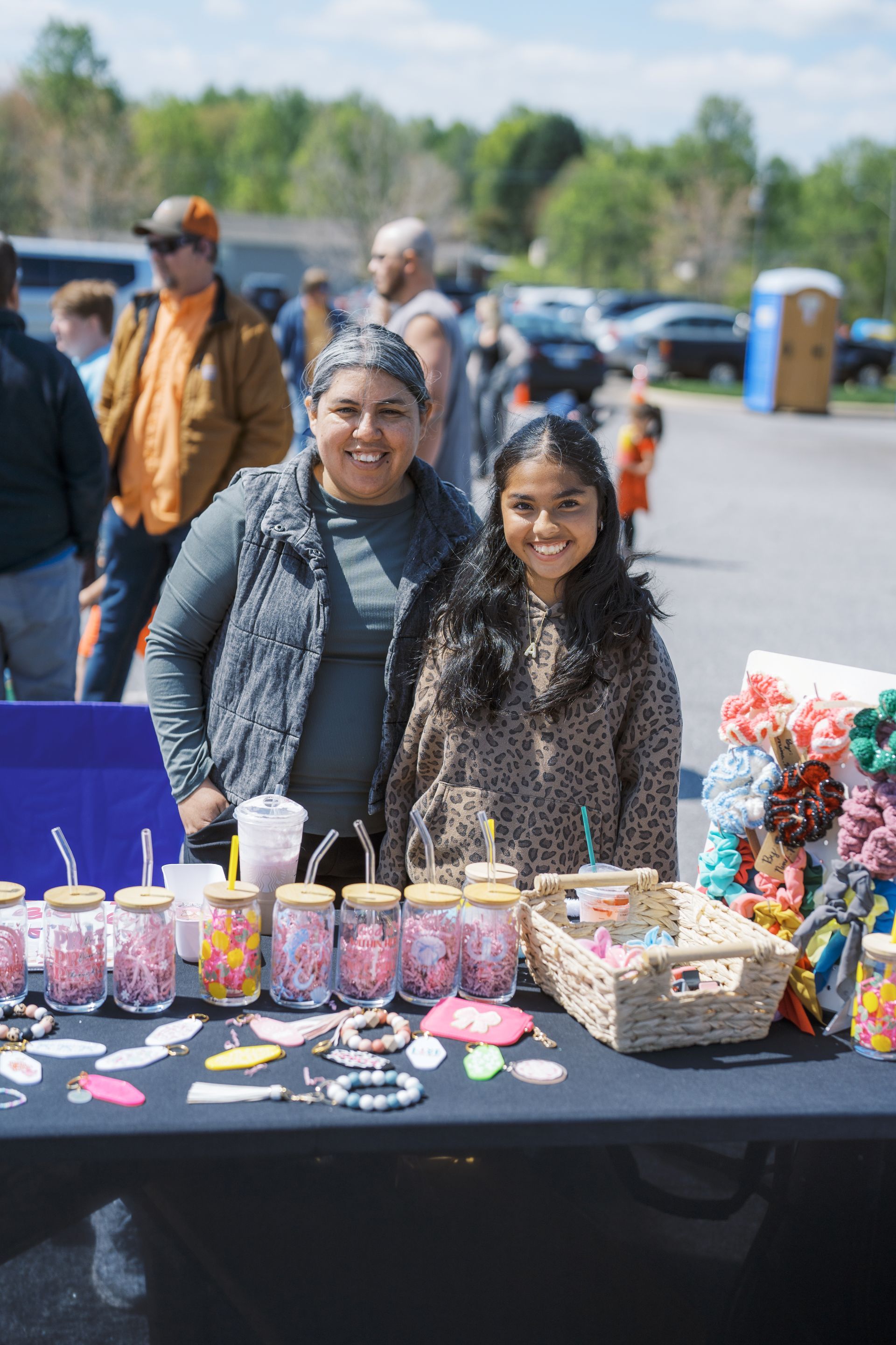 Two vendors smiling behind a craft table with jewelry, candles, and small baskets outdoors