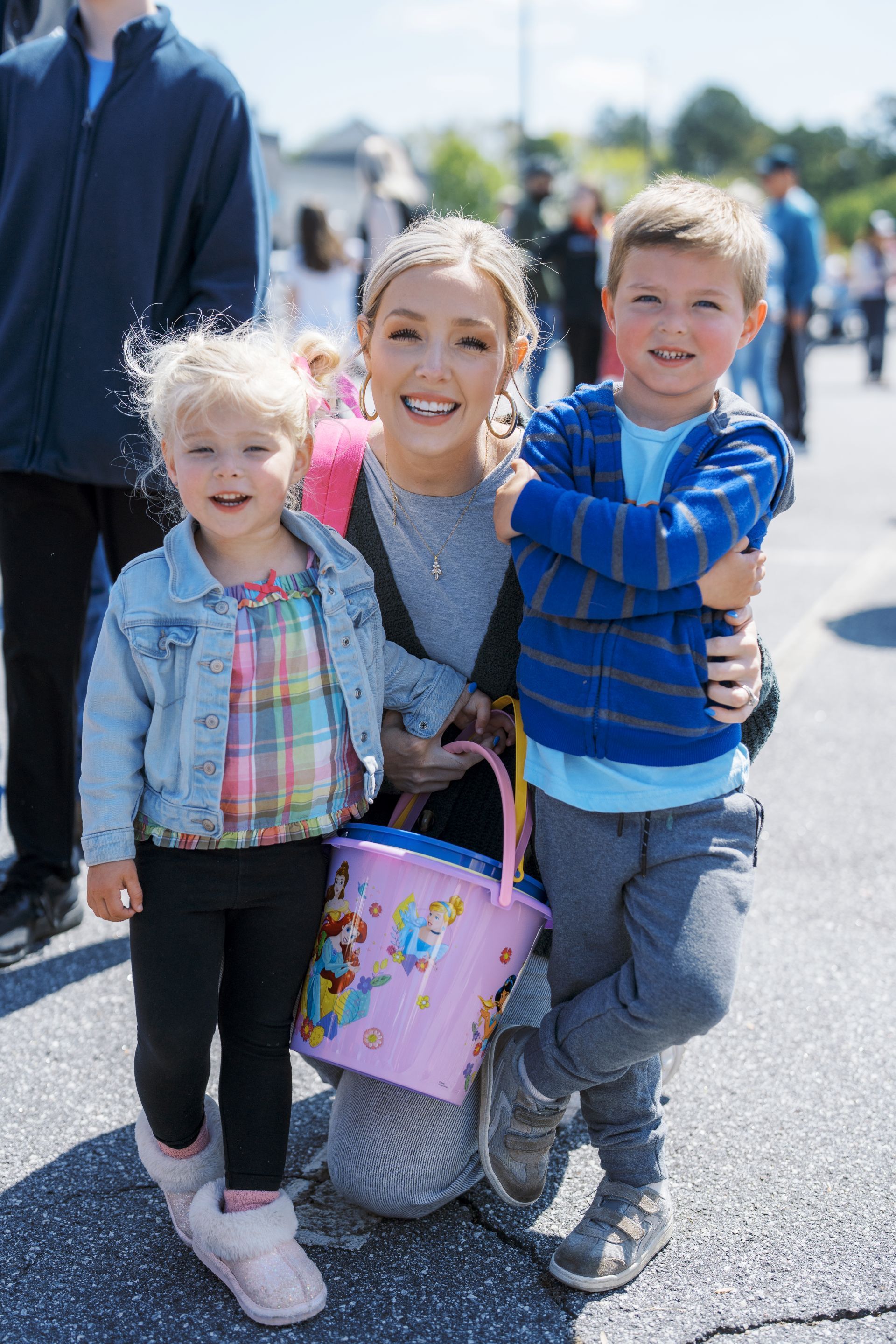 Smiling woman kneels between two children outdoors, holding a pastel bucket on a sunny street.