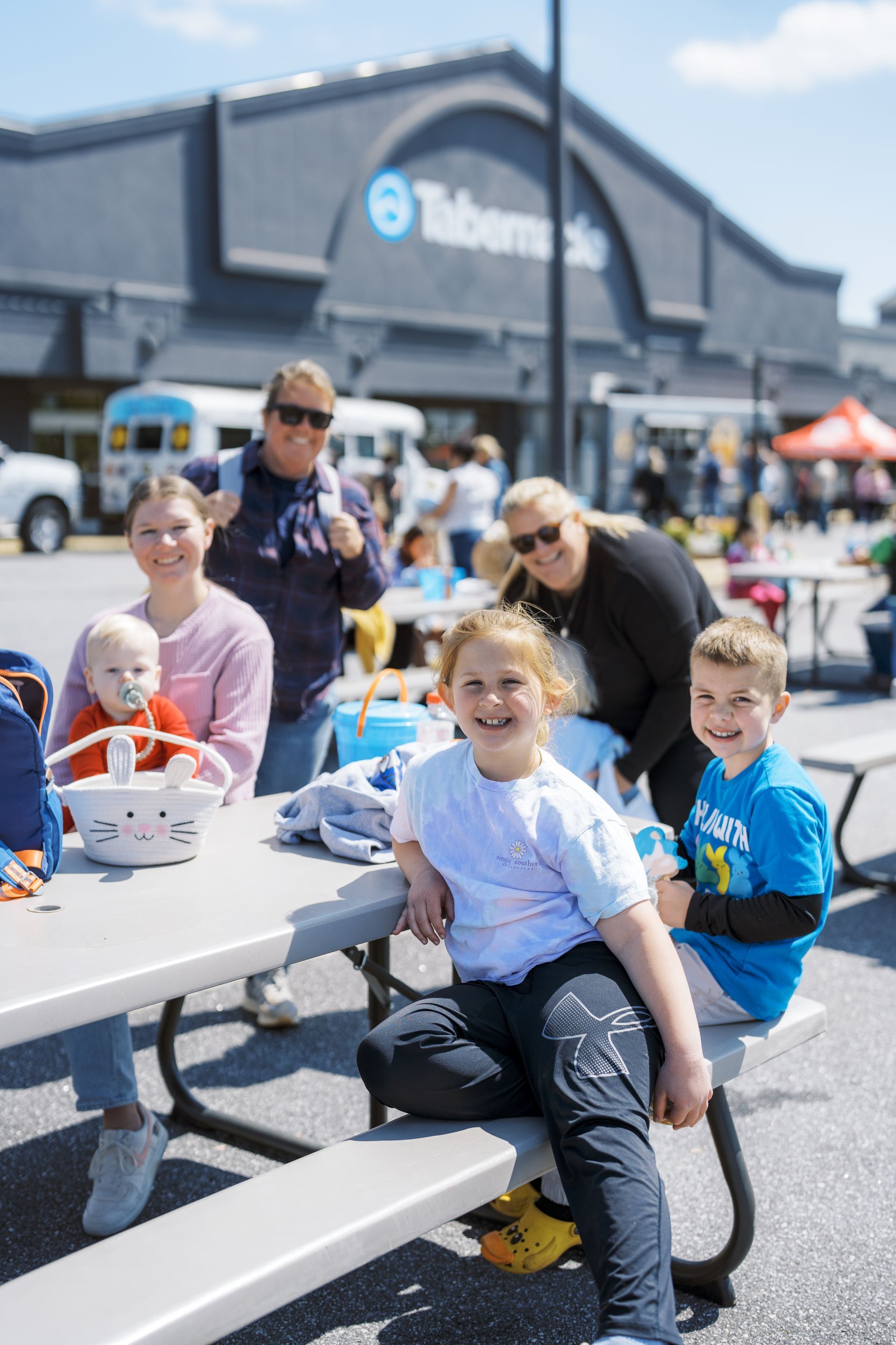 Children and adults seated at picnic tables outside a T-Mobile store, smiling on a sunny day