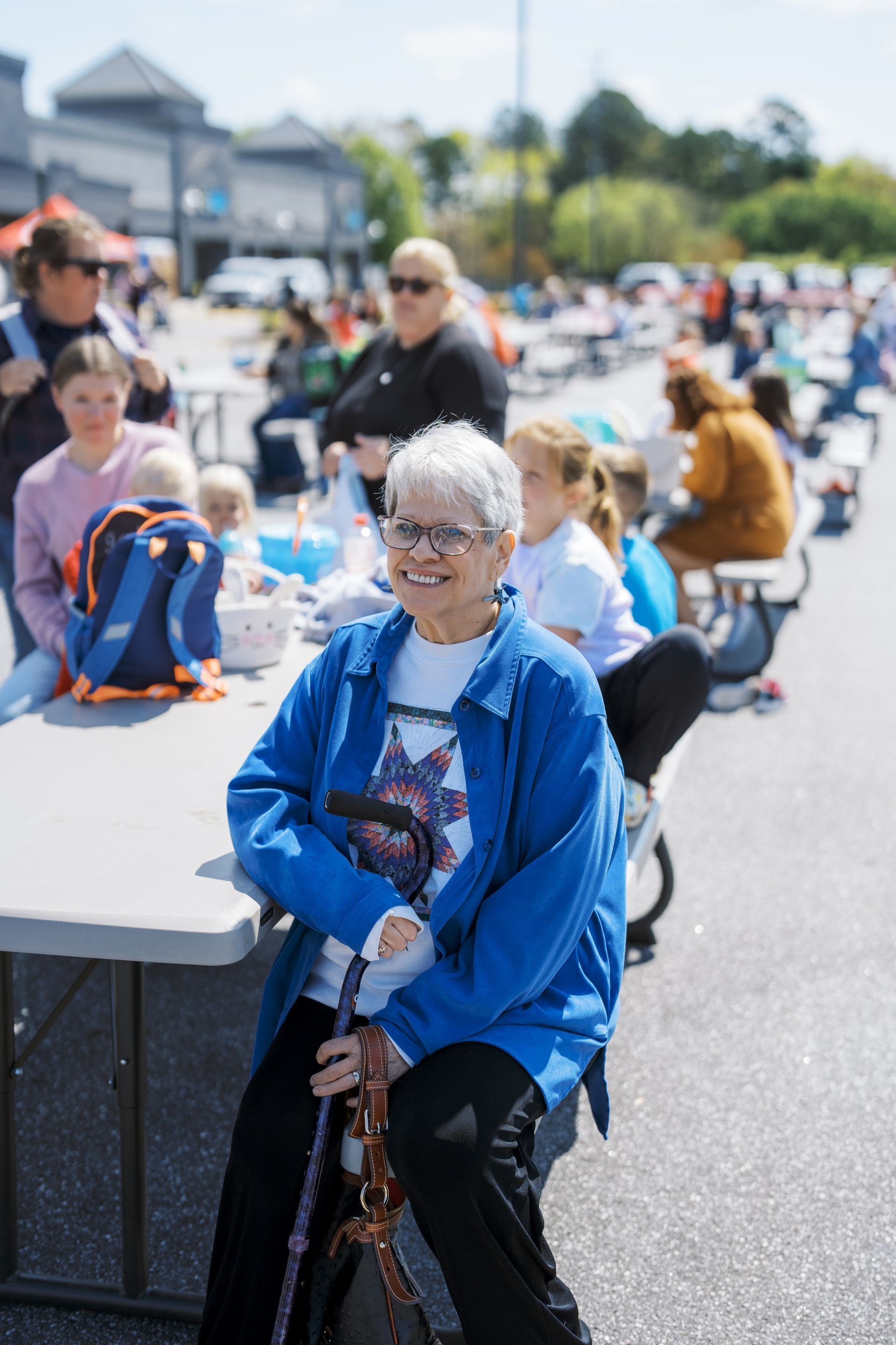 Smiling woman in blue jacket sitting at an outdoor table with people and bicycles in the background