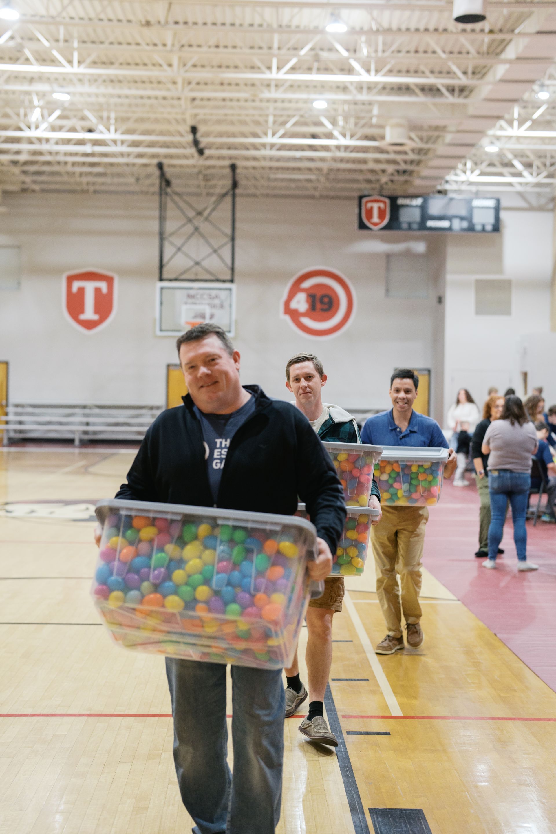 People carrying clear bins of colorful balls in a gymnasium with bleachers and hanging signs