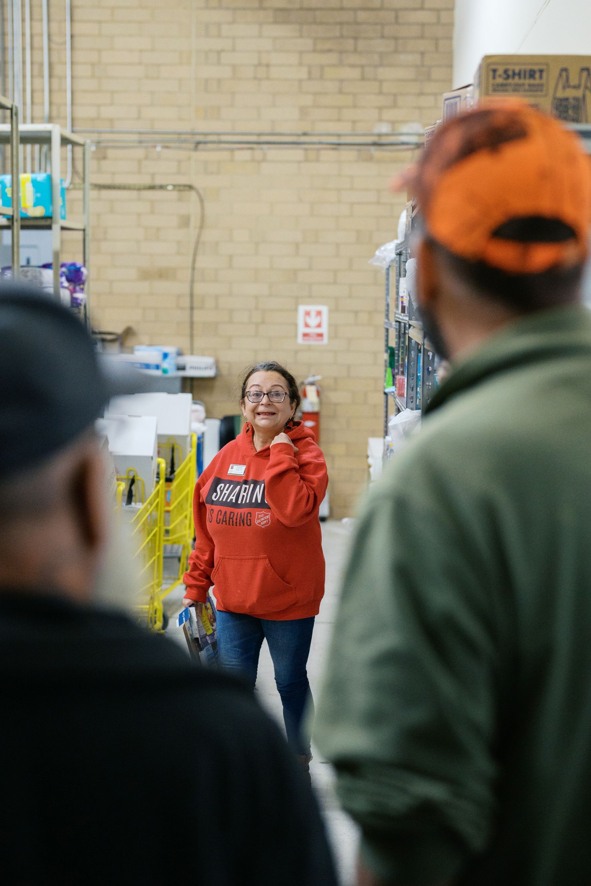 Person in a red hoodie smiling in a warehouse aisle, with two blurred people in the foreground.