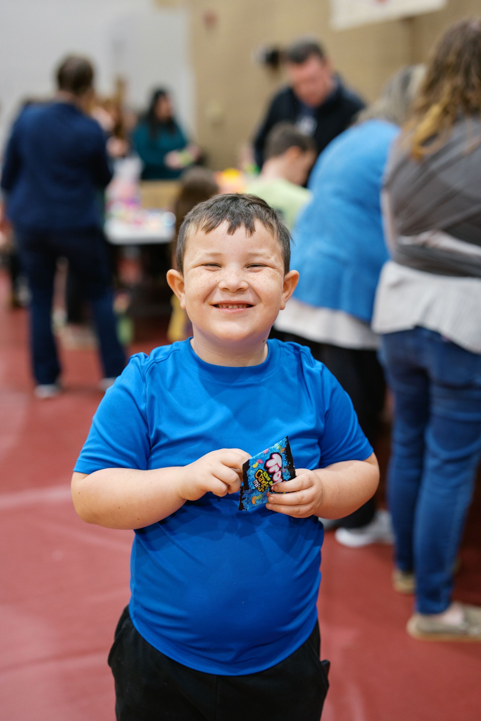Smiling child in a blue shirt holding a small toy at an indoor event with people blurred in the background