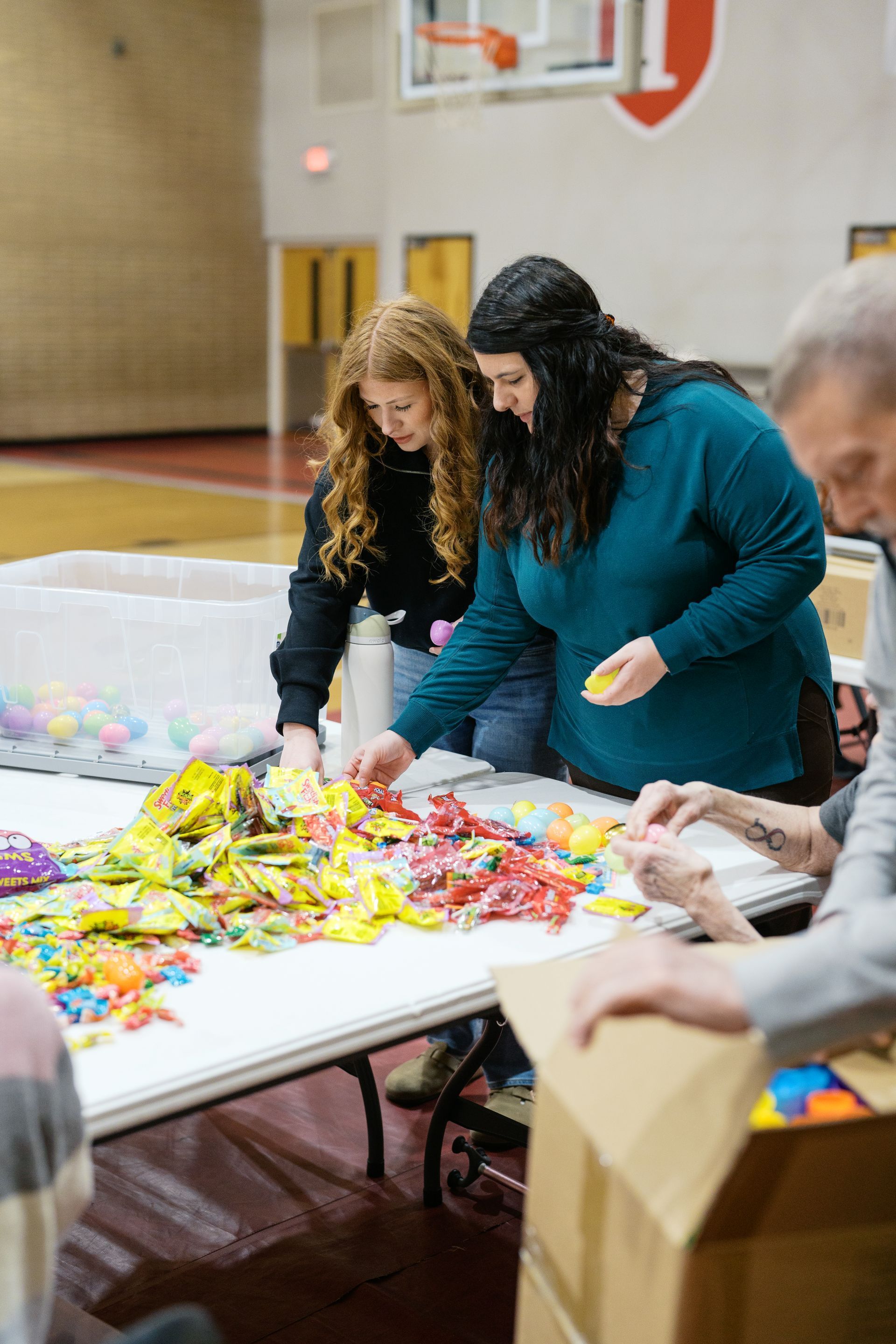 Two women sorting colorful packaged items at a table in a gymnasium