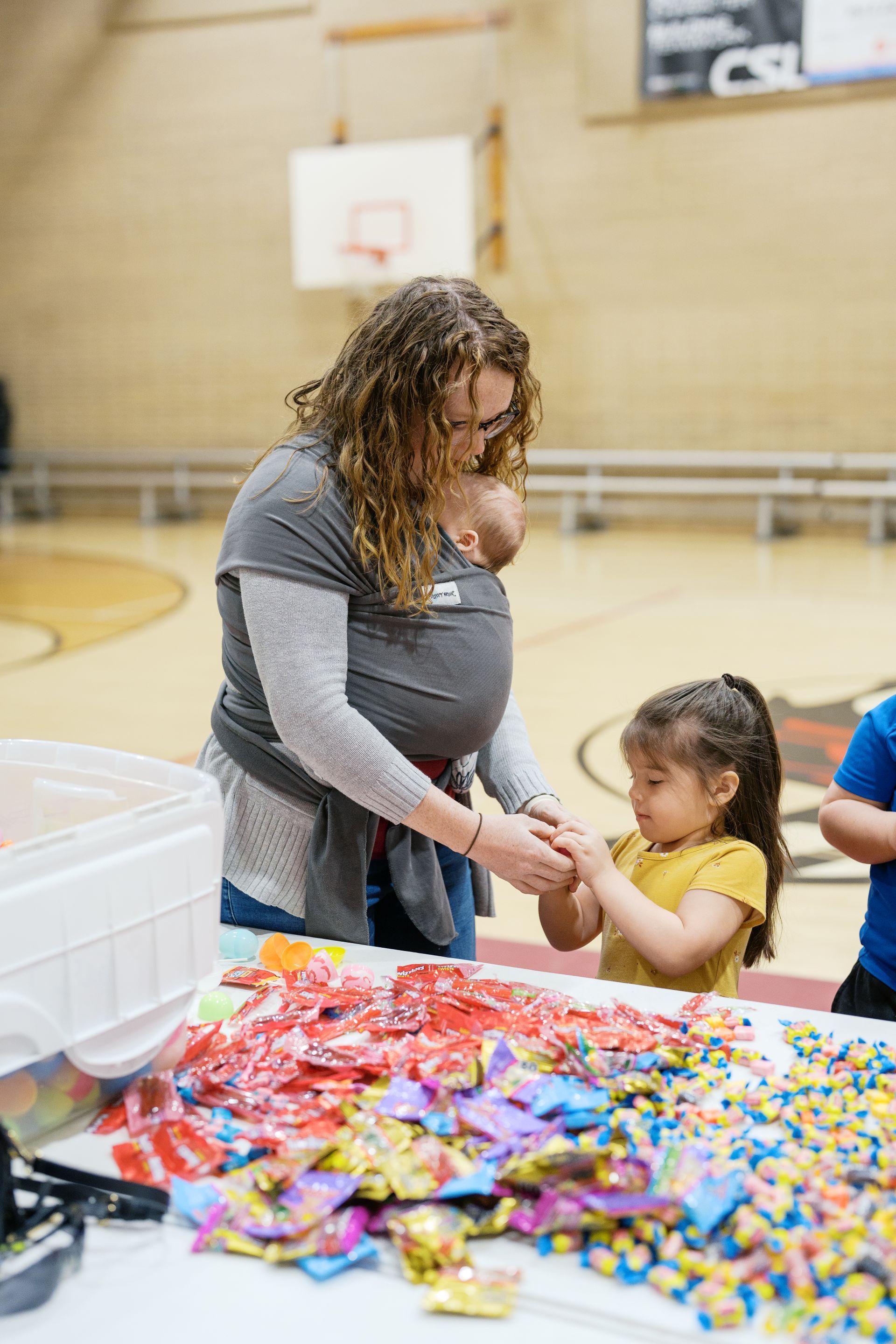 Woman helping a child with a craft activity at a table full of colorful candy in a gymnasium