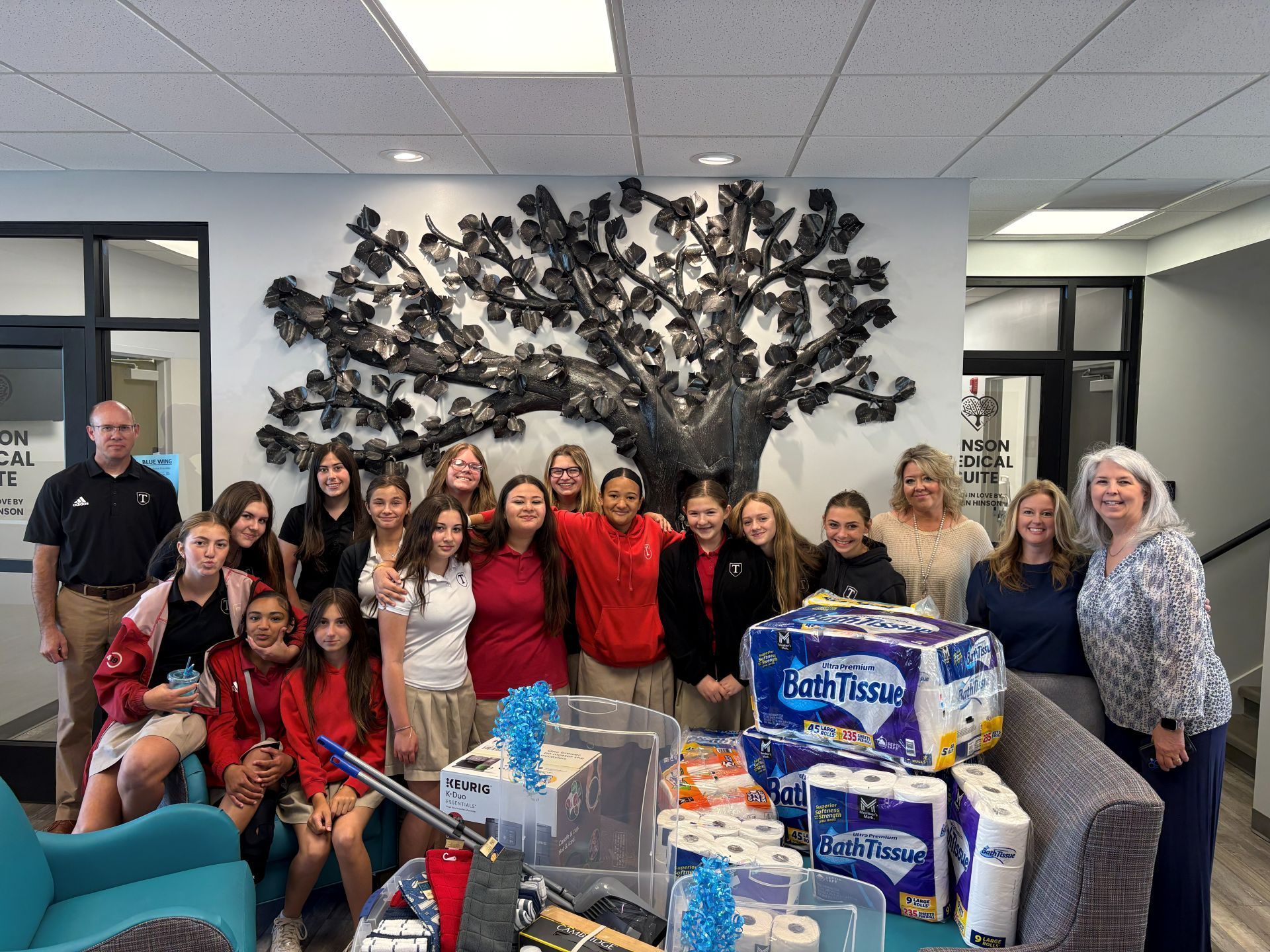 Group posing indoors with donated supplies and cases of water in front of a wall sculpture.