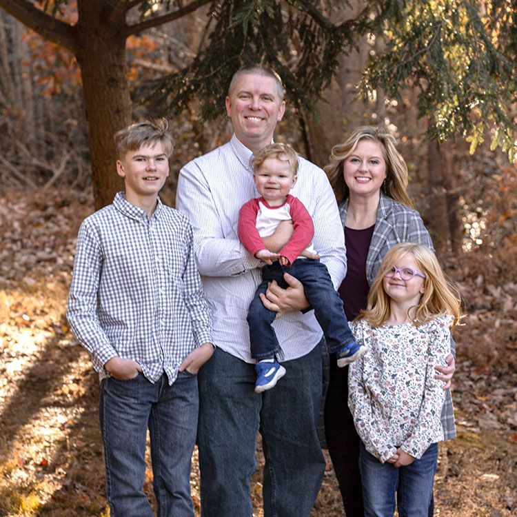 Family of five standing outdoors in a wooded area, with three children and two adults smiling at the camera.