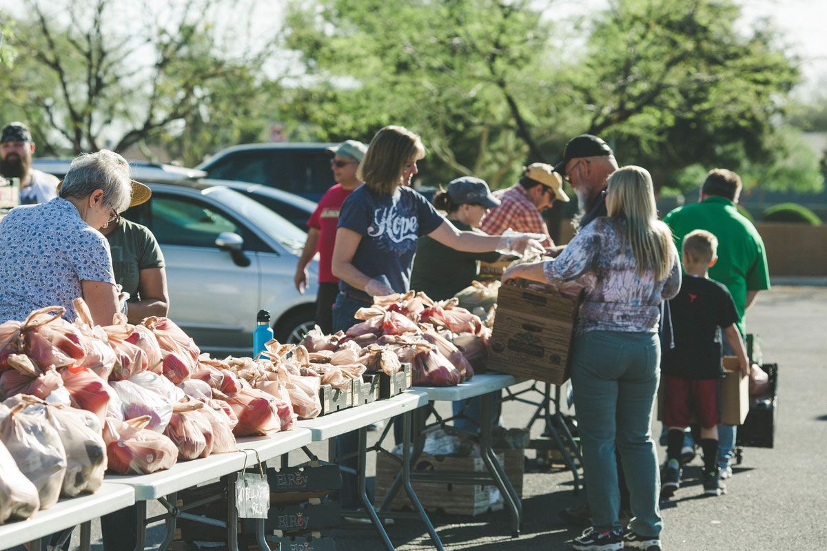 People browsing a roadside market table piled with meat packages, with cars parked behind them.