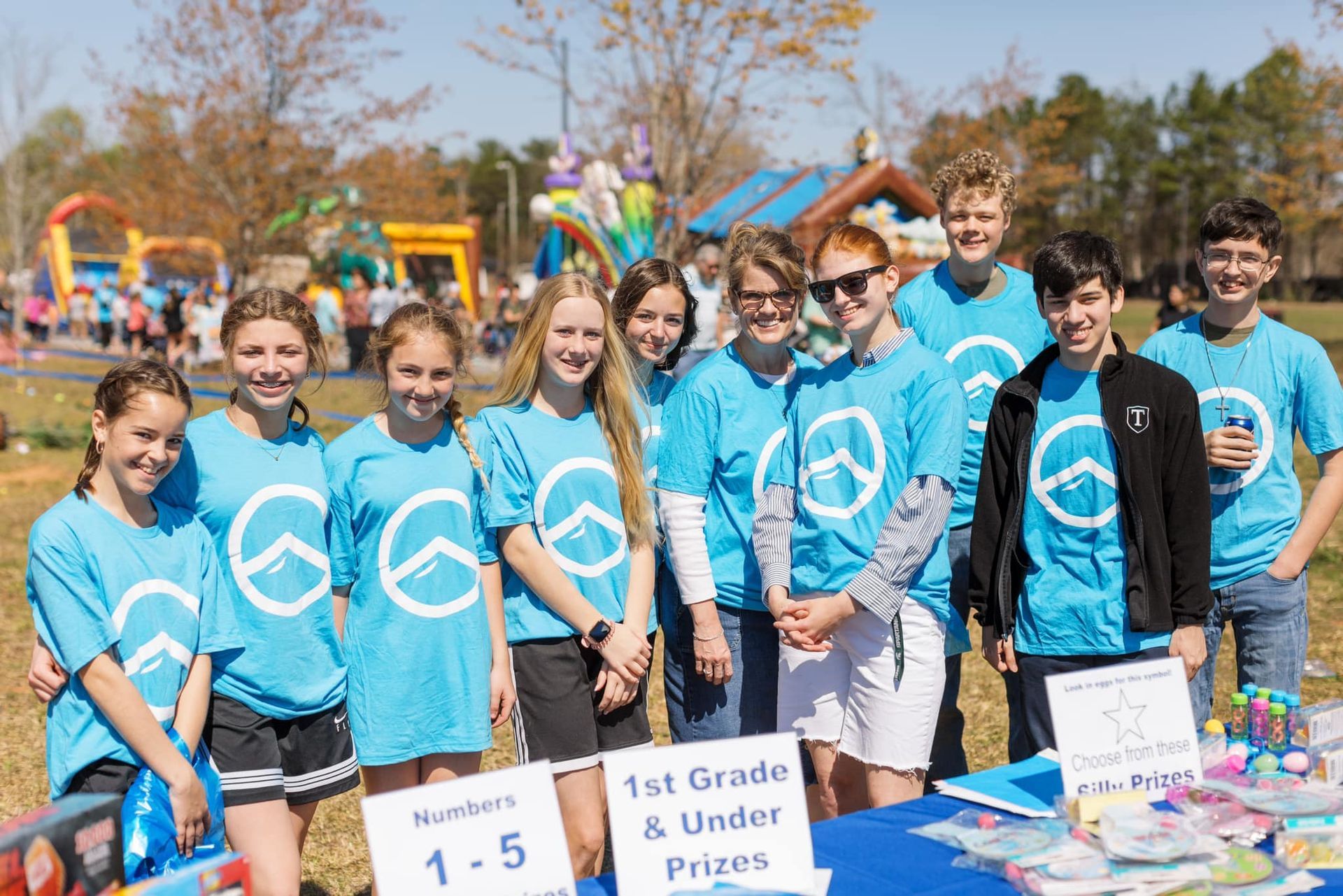 Group in blue shirts posing behind “1st Grade & Under” sign at an outdoor event.