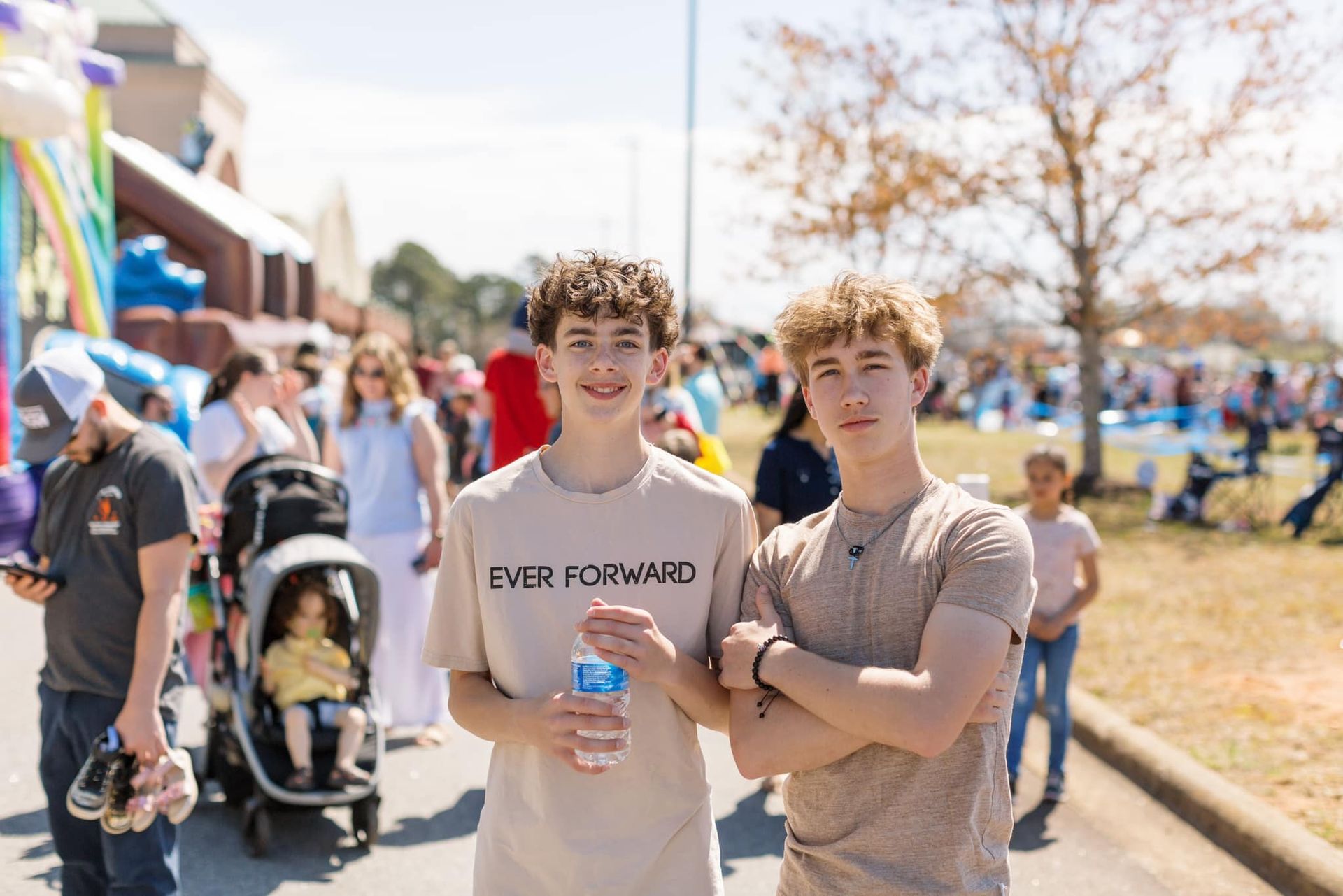 Two smiling people stand at a crowded outdoor fair, one holding a water bottle.