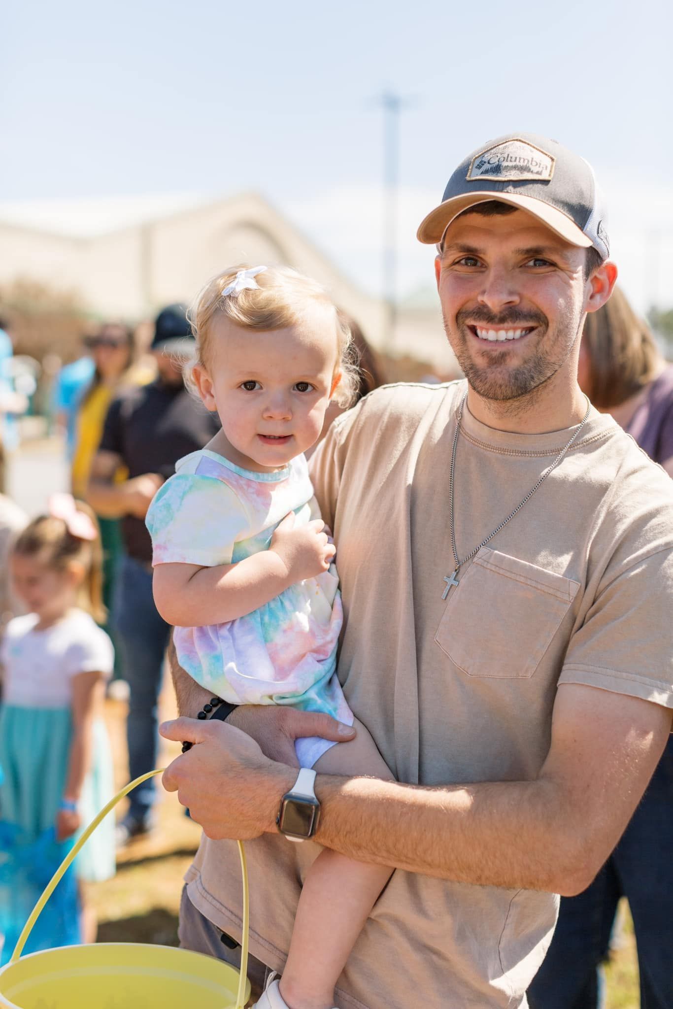 Man holding a smiling toddler outdoors in bright sunlight at a festival