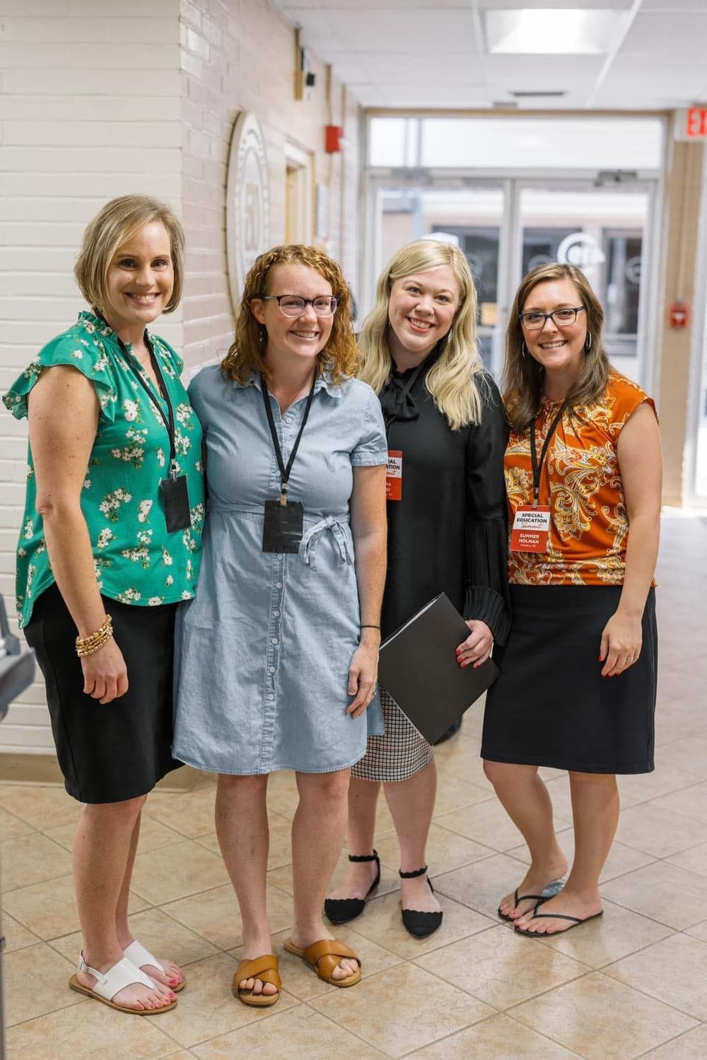Four women stand smiling in a hallway, wearing business-casual outfits.