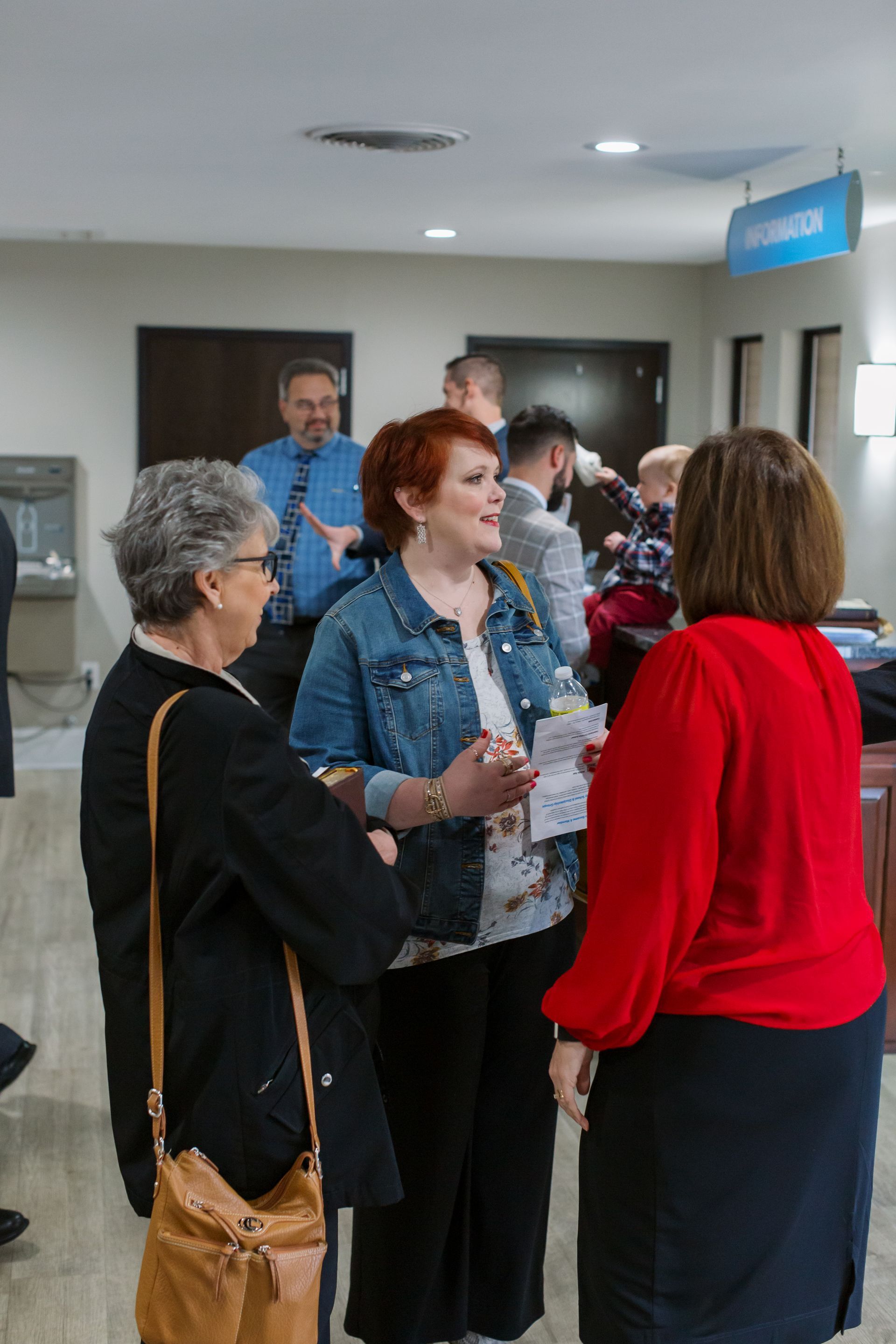 People mingling at an indoor event, chatting near a hallway with a blue sign overhead.
