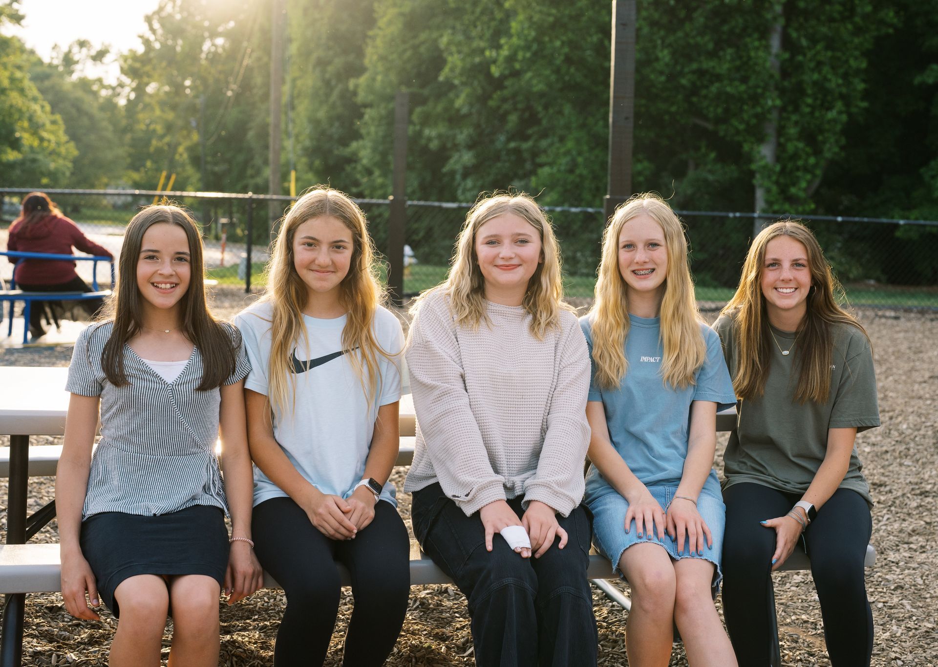 Five smiling people sitting on a bench outdoors in sunlight, with trees and a fence behind them.